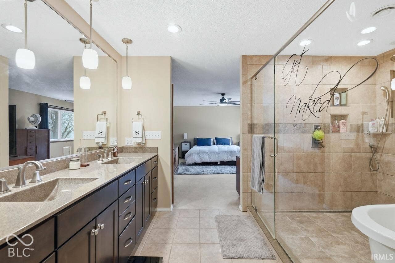 Double vanity with dark wood cabinetry, solid-surface countertops, dual integrated sinks, glass-enclosed shower with tile surround, and recessed lighting