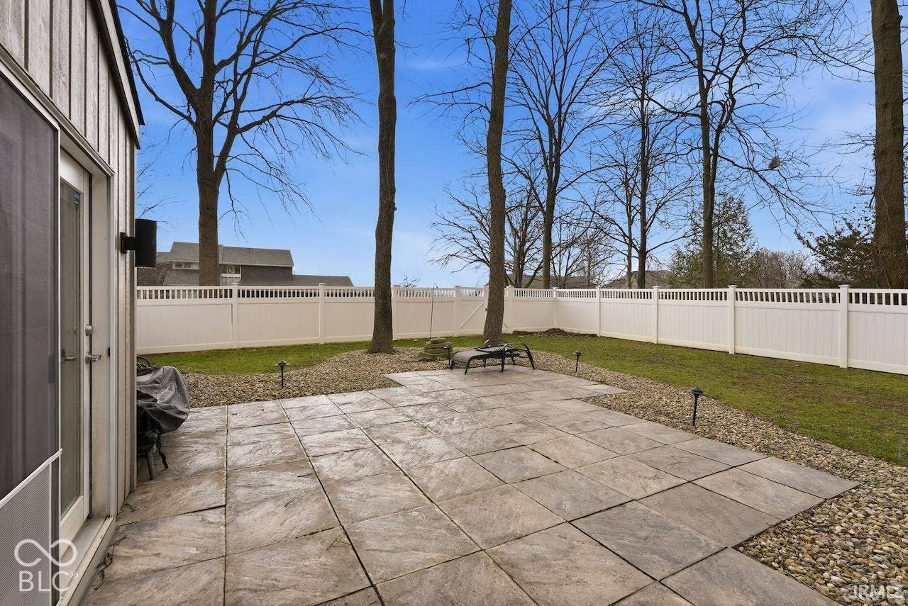 Rear exterior featuring a large-format tiled patio, stone-accented landscape borders, white vinyl privacy fencing, and mature trees