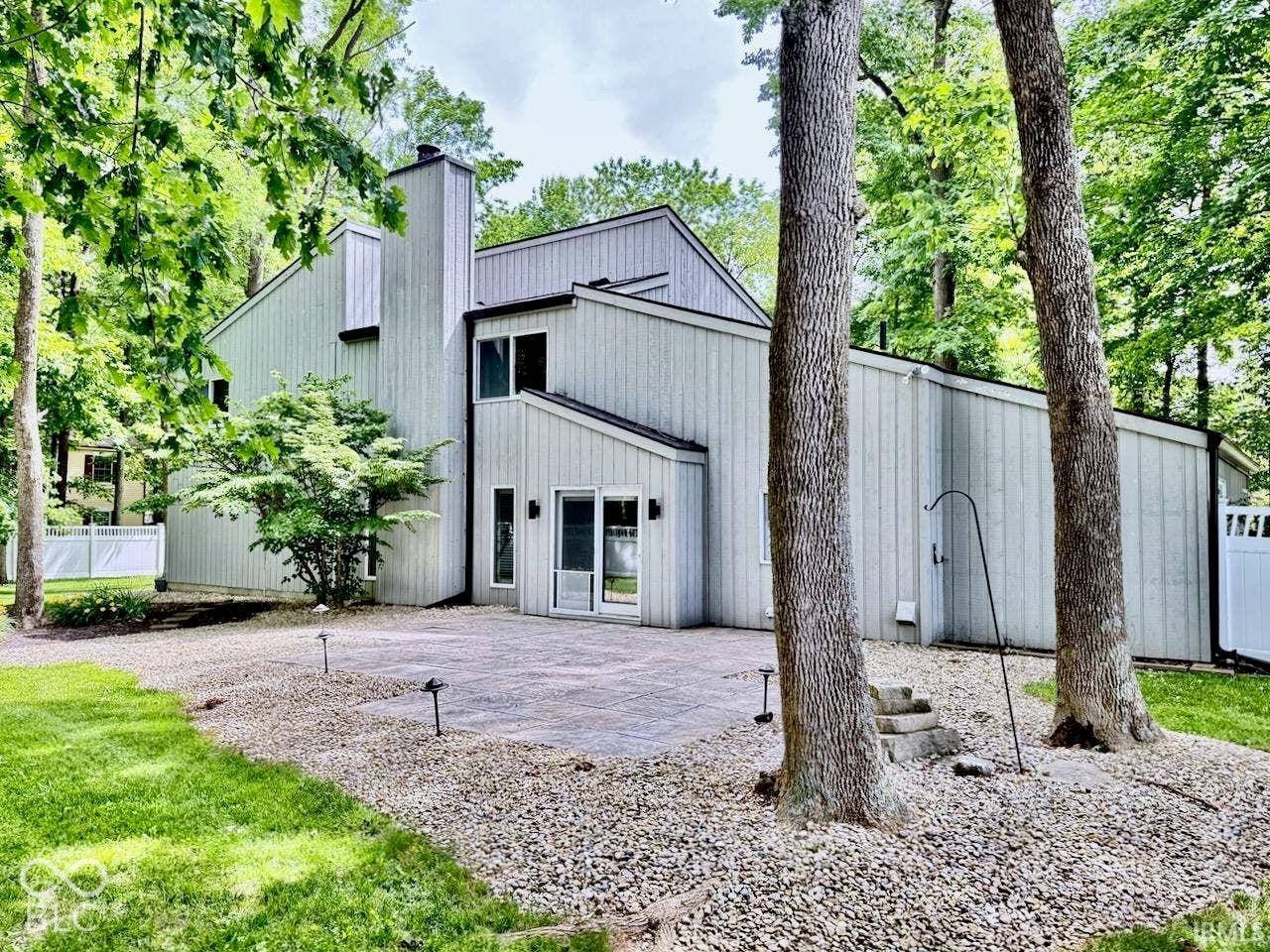 Vertical wood siding exterior featuring a multi-level roofline, prominent exterior chimney, stone-paver patio, and mature trees