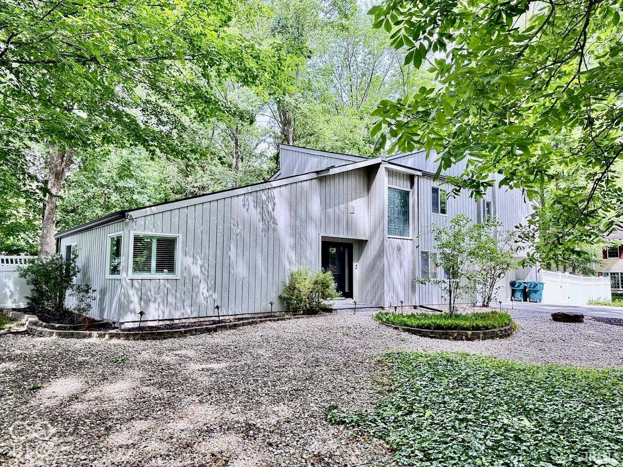 Vertical wood-paneled siding, multifaceted roofline, multi-pane windows, and stone-bordered landscaping situated on a gravel driveway
