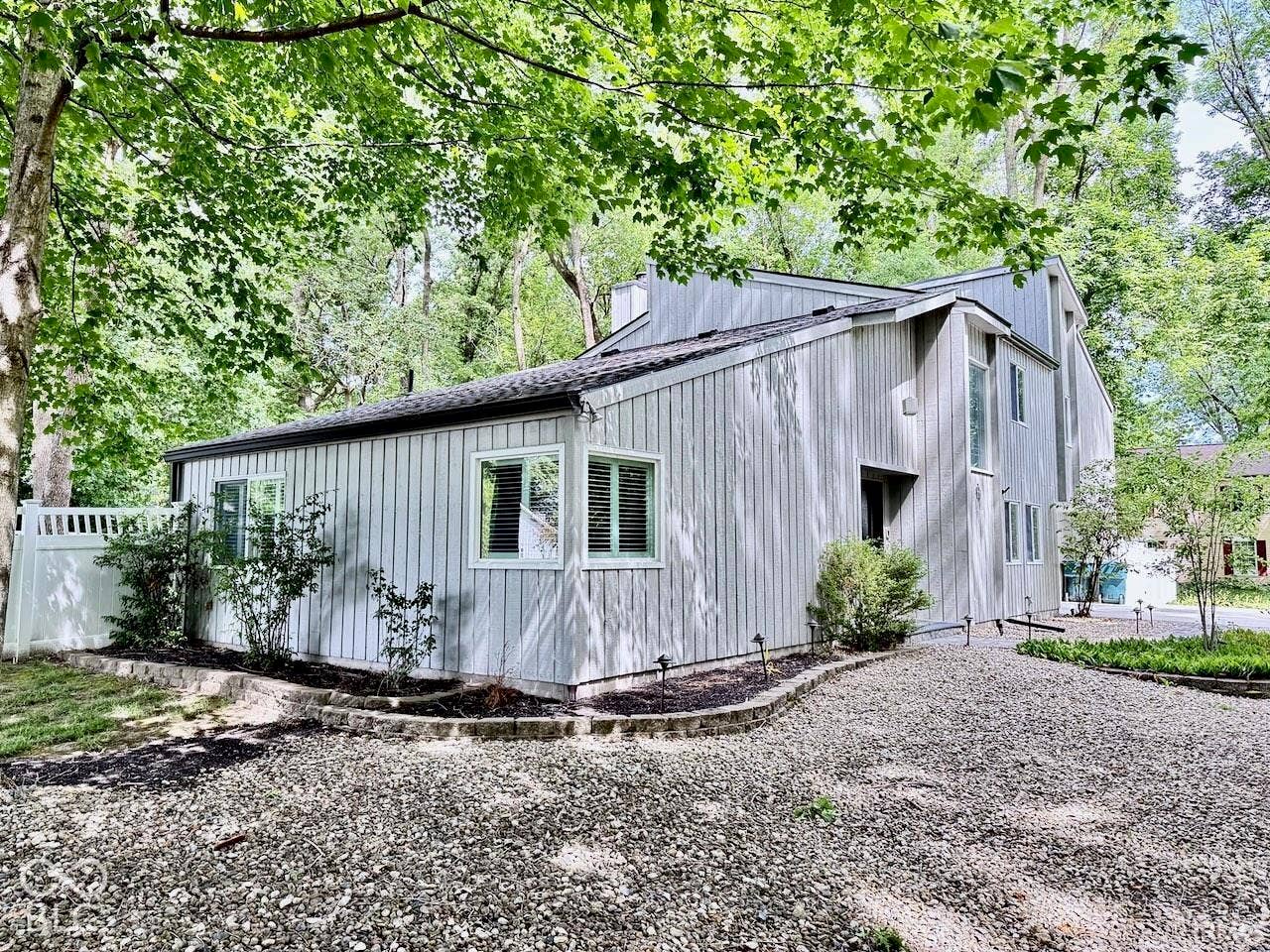 Multi-level exterior featuring vertical wood-finish siding, stone garden bed borders, a gravel driveway, and a white vinyl privacy fence