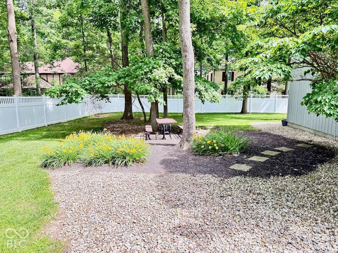 Private backyard featuring a white vinyl privacy fence, mature trees, landscaped garden beds with perennials, and stone path accents