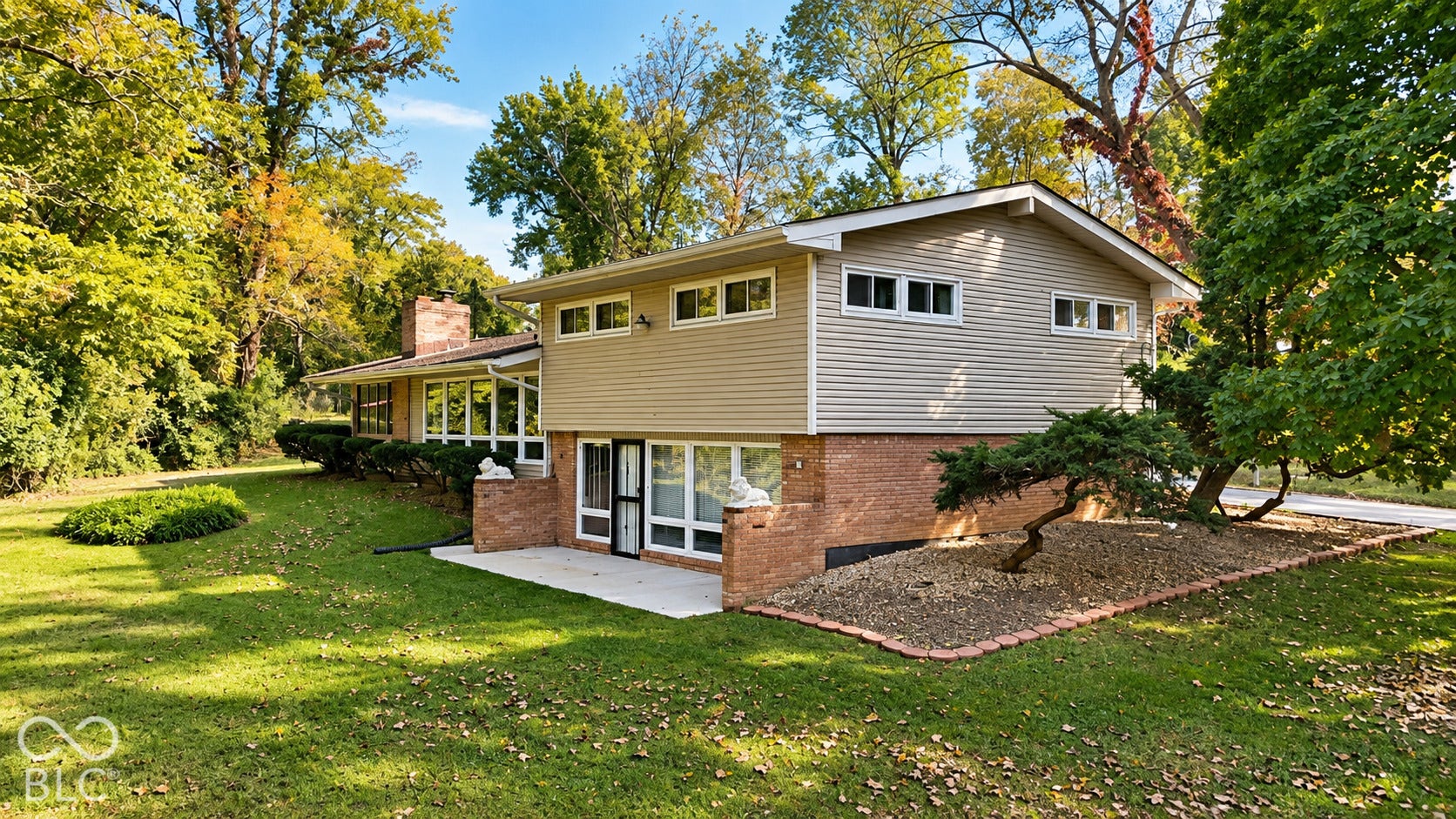 4225 Westbourne Drive, Indianapolis, IN 46205 Split-level exterior featuring light-toned horizontal siding, a lower-level brick facade, a masonry chimney, a concrete patio, and landscaped beds with stone ground cover