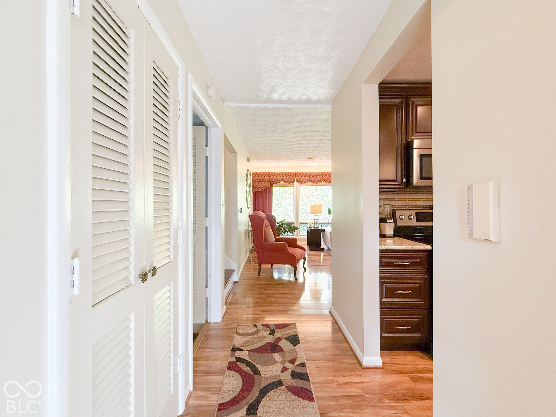 4225 Westbourne Drive, Indianapolis, IN 46205 Hallway featuring wood-finish flooring, white louvered-style bi-fold closet doors, and recessed lighting, with visibility into a kitchen area containing dark-stained cabinetry and stone-finish countertops
