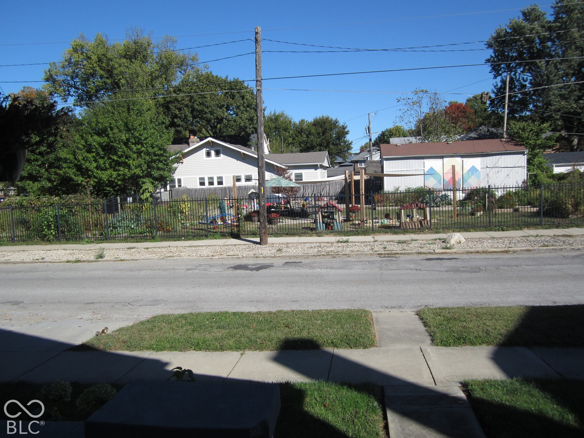 Fenced exterior lot featuring mature trees, landscaped planting beds, and a concrete sidewalk