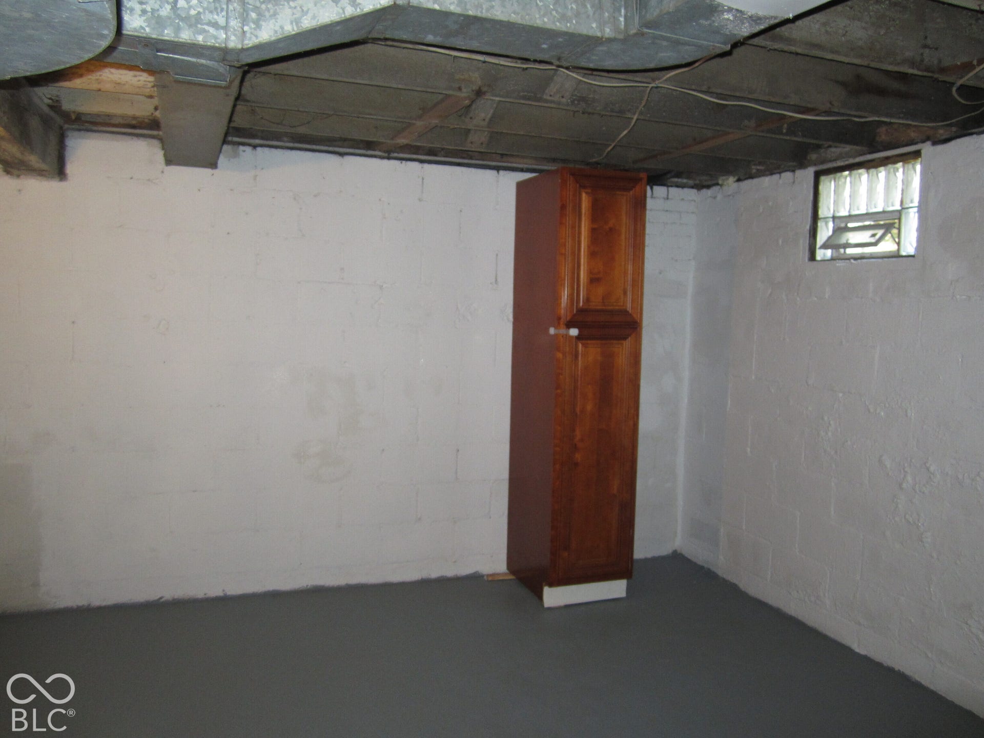 Unfinished space featuring painted concrete block walls, a solid-color finished floor, exposed wood ceiling joists, and a built-in wood-finish storage cabinet with an inset glass block window