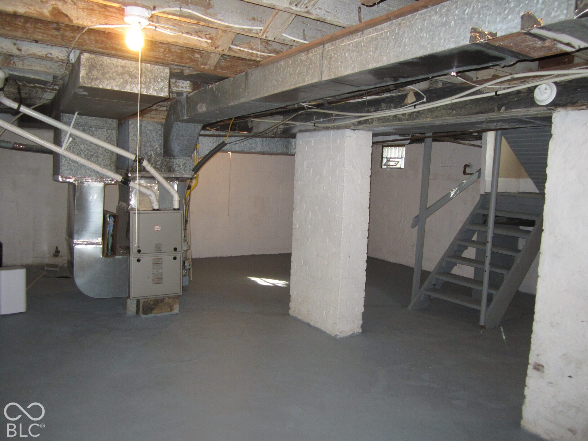 Unfinished lower level featuring painted concrete flooring, structural masonry support pillars, metal staircase, and exposed ceiling joists with metallic ductwork