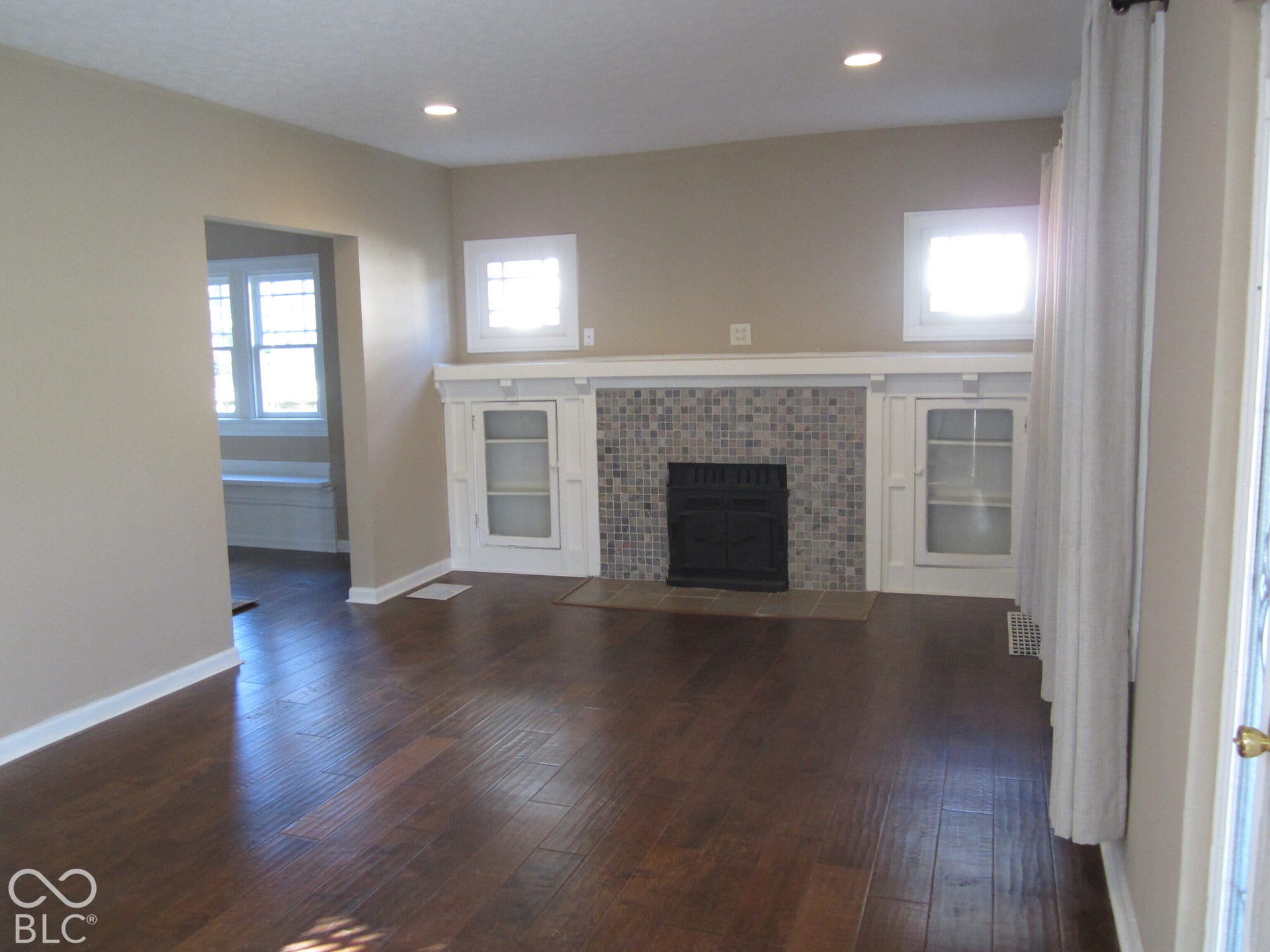 Wood-finish flooring throughout, featuring a fireplace with tiled surround, built-in cabinetry with glass-front doors, recessed lighting, and dual square windows