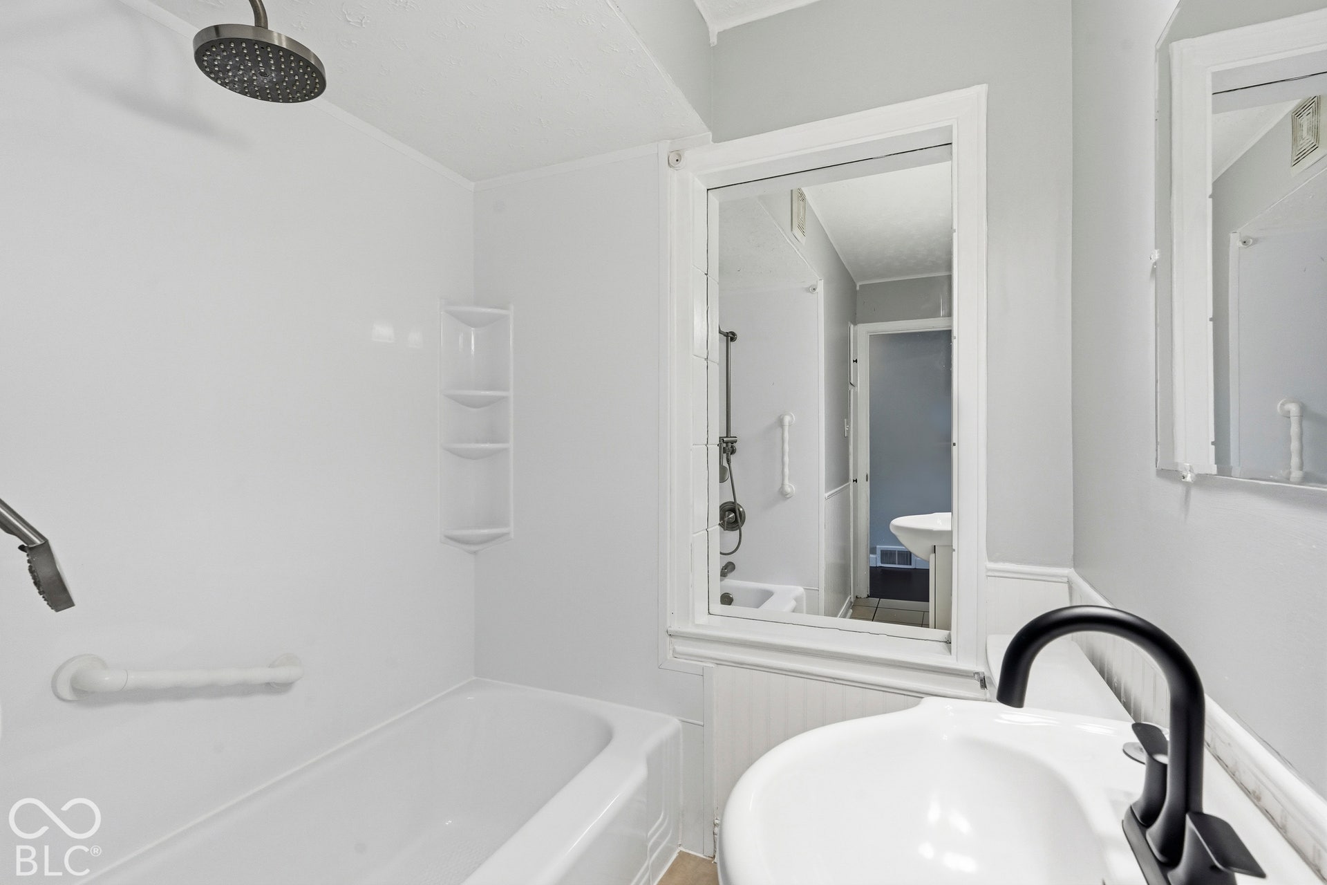 Bathroom featuring a white bathtub with integrated shelving, a round rain-style shower head, a pedestal sink with a matte black faucet, and a wall-mounted mirror