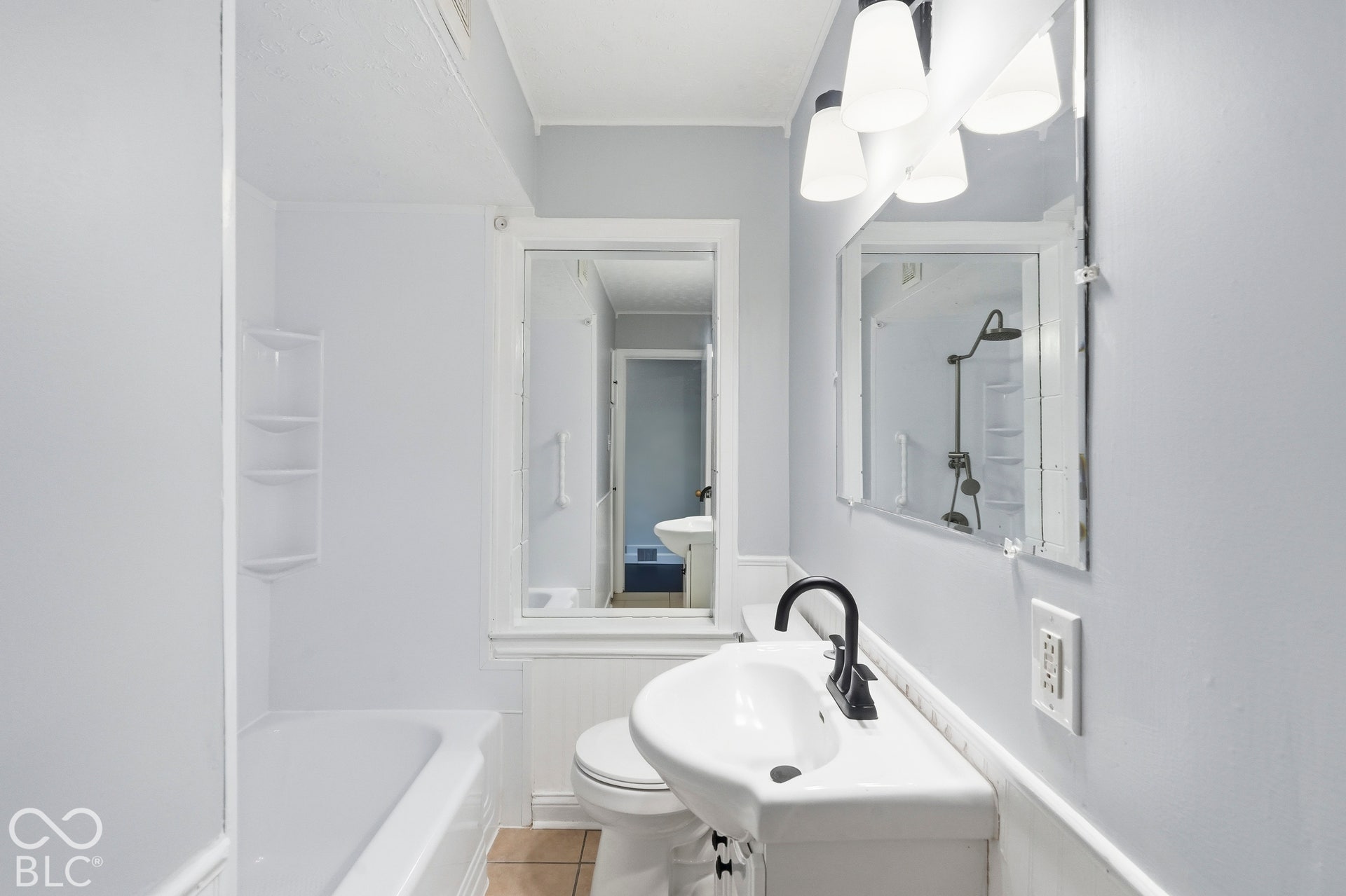 Bathroom featuring a pedestal sink with a matte black faucet, wall-mounted mirror with three-bulb light fixture, and a tub-shower combination with integrated shelving