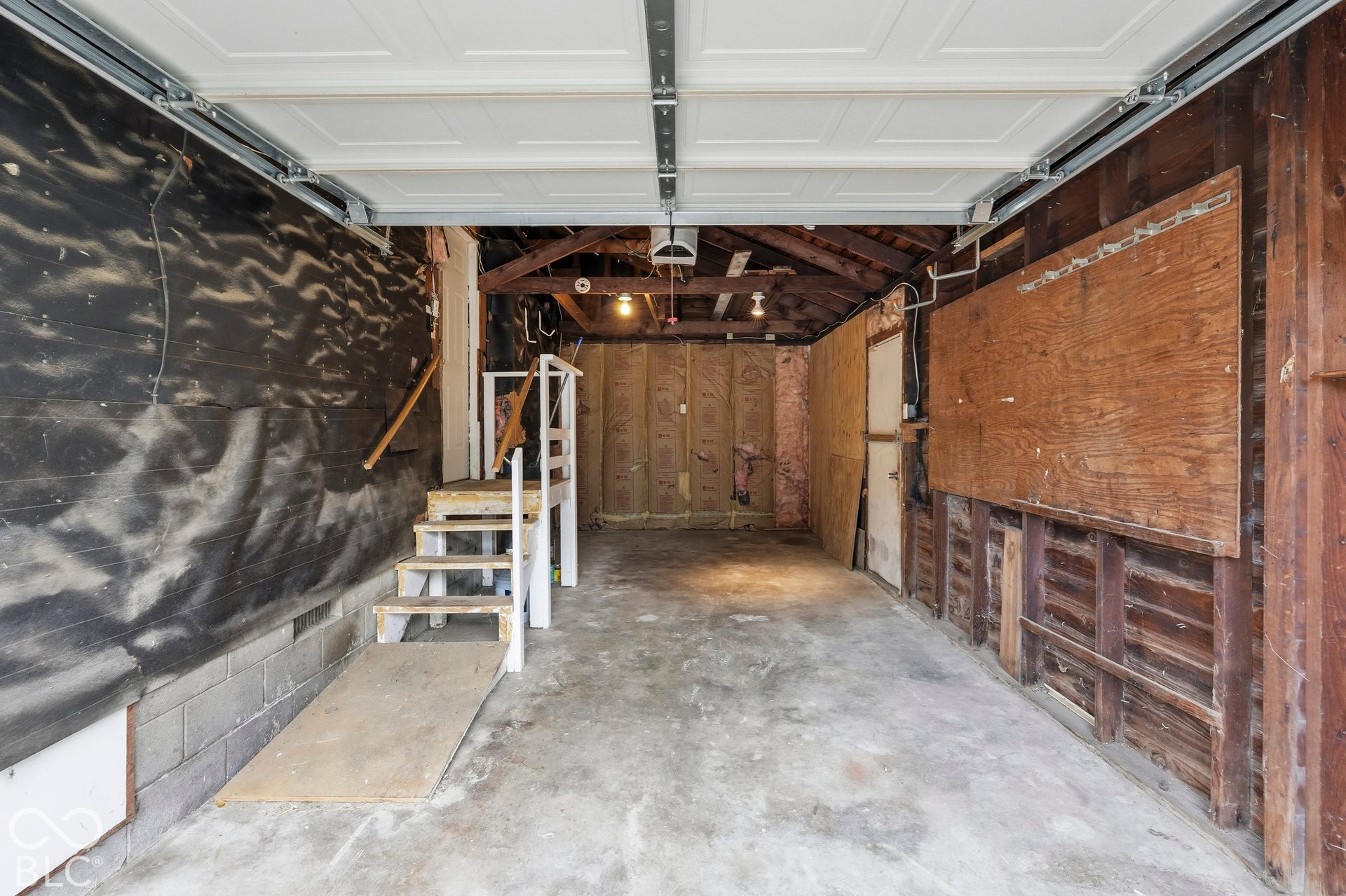 Enclosed garage featuring a concrete floor, sectional overhead door, exposed wood-beam ceiling, and integrated wooden staircase