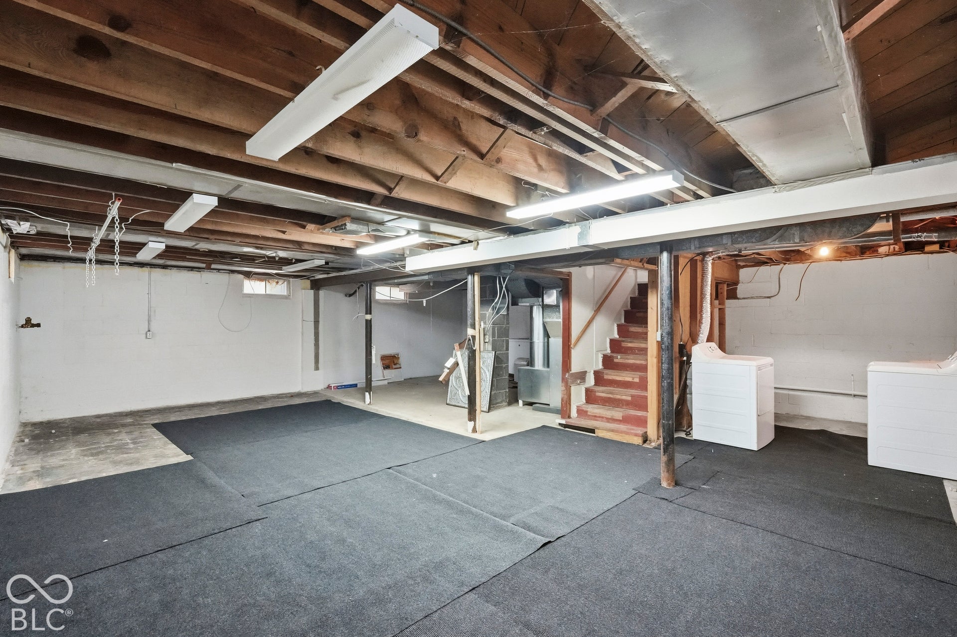 Open-concept lower level featuring exposed wood joist ceilings, integrated linear fluorescent lighting, dark carpeting, and structural support columns