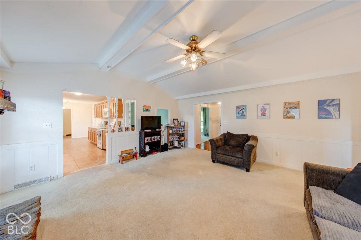 Living space featuring vaulted ceilings with exposed beams, a central ceiling fan with integrated lighting, and white wainscoting