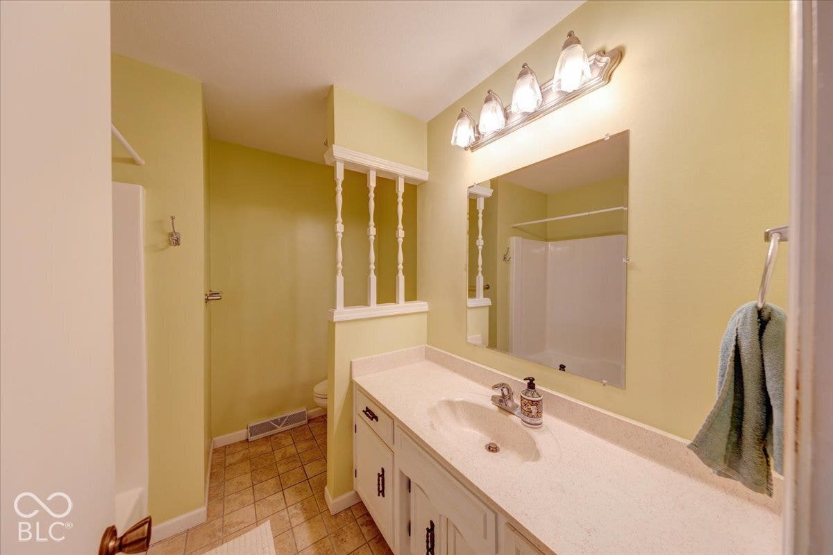 Vanity featuring a light-toned solid surface countertop with an integrated oval sink, four-light vanity fixture, wall-mounted mirror, white cabinetry, and a decorative spindle partition