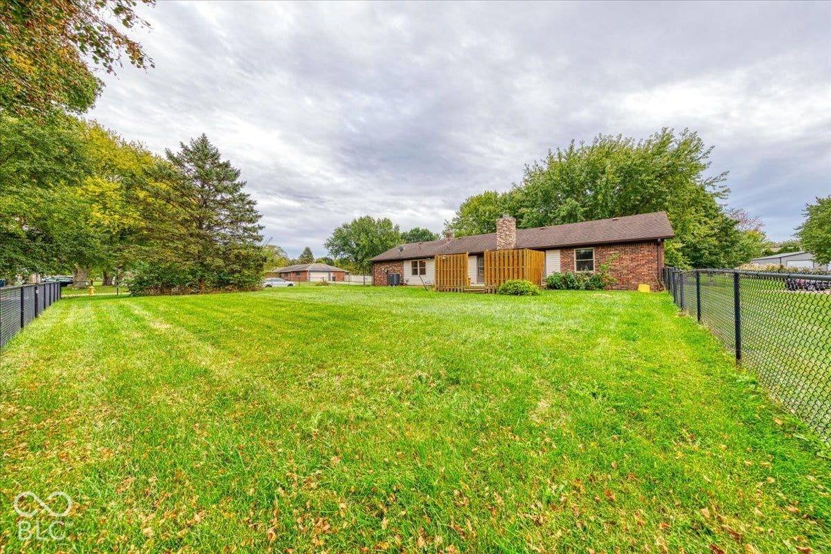 Expansive backyard featuring a level lawn, mature trees, chain-link perimeter fencing, and a brick exterior house with a stone chimney