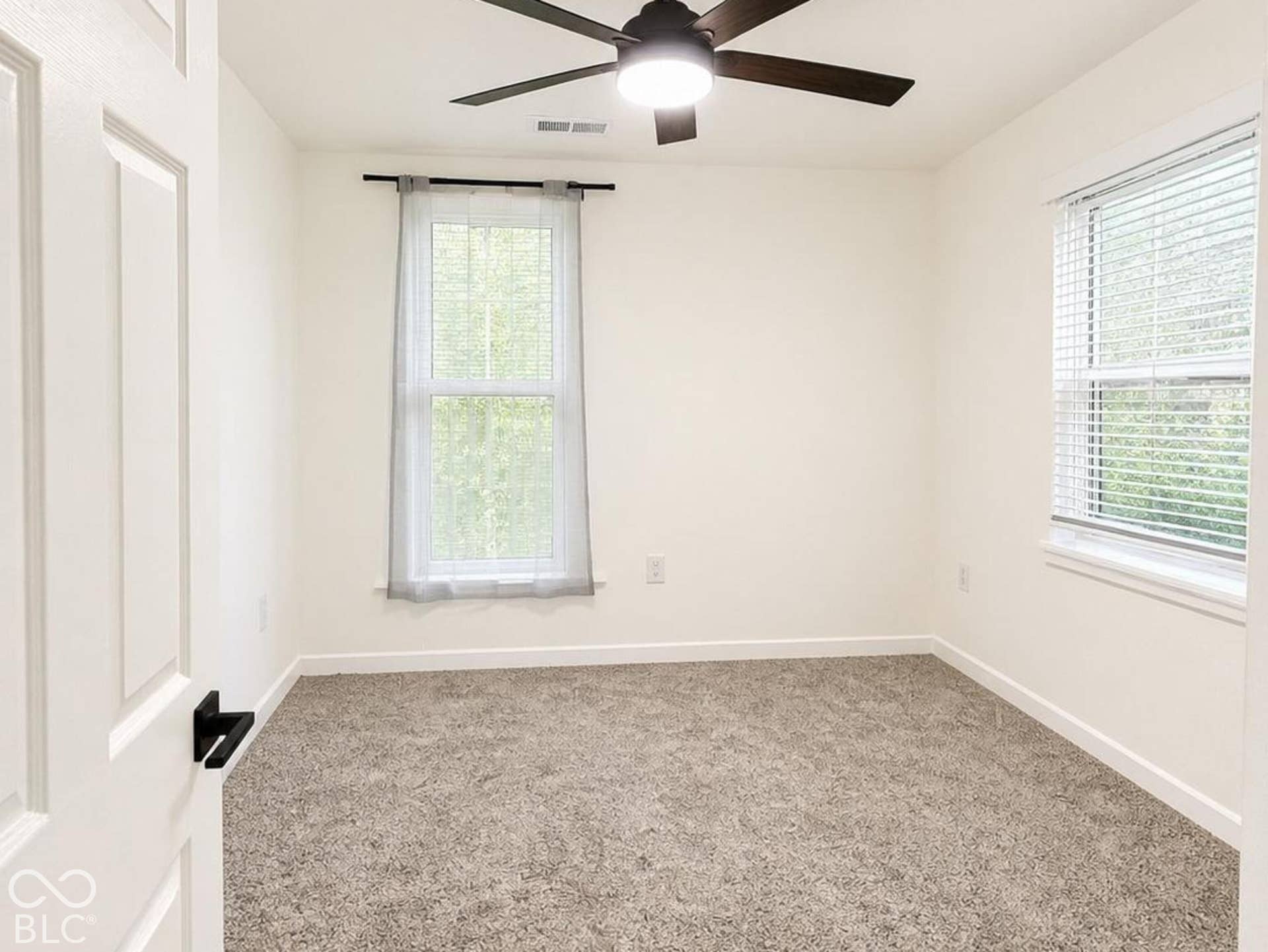 Neutral-toned carpeting, five-blade matte black ceiling fan with integrated lighting, dual windows with blinds, and solid-core interior door with matte black hardware