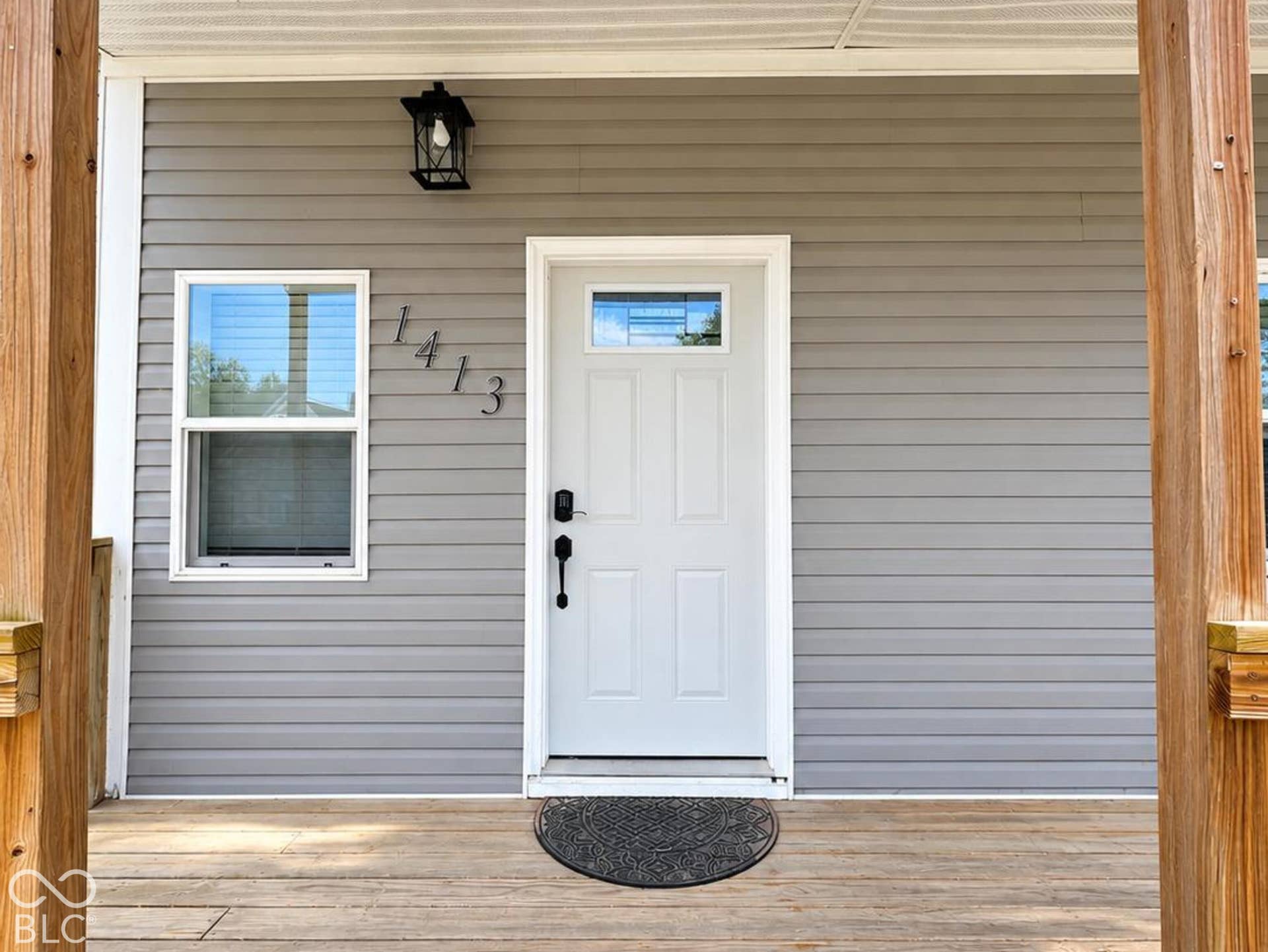 Covered porch featuring horizontal wood-finish decking, gray horizontal siding, a white front door with an integrated window, and a wall-mounted lantern-style light fixture