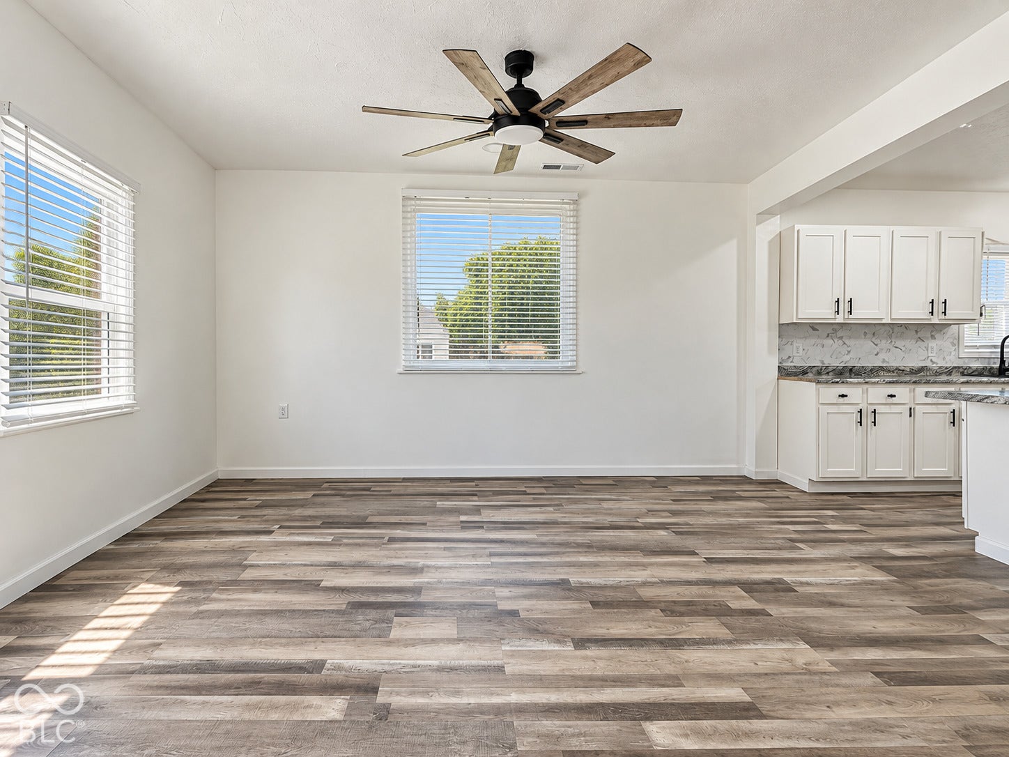 Wood-finish flooring, multi-blade ceiling fan with integrated lighting, white cabinetry, and stone-patterned backsplash