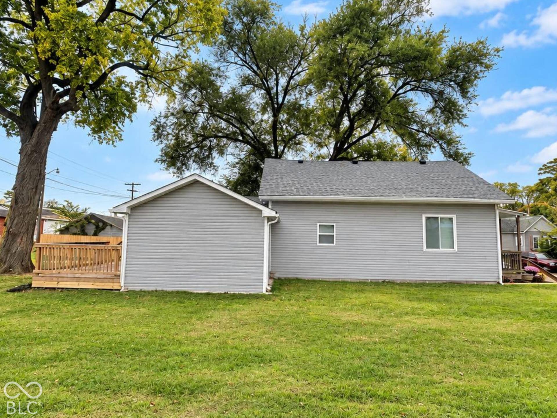 Exterior featuring grey horizontal siding, asphalt shingle roofing, and an attached wood deck set upon an expansive lawn with mature trees