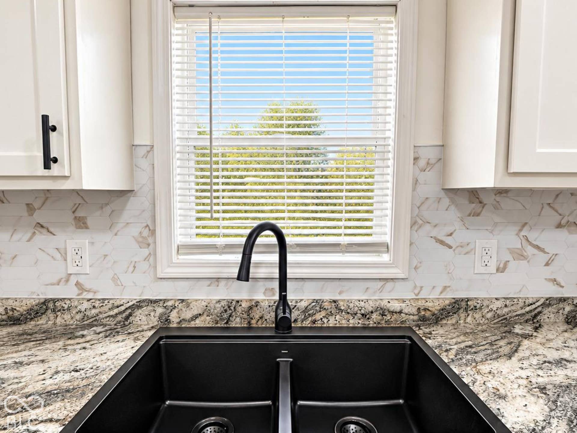 Dual-basin black composite sink with a high-arch matte black faucet, stone-finish countertops, patterned tile backsplash, and shaker-style white cabinetry