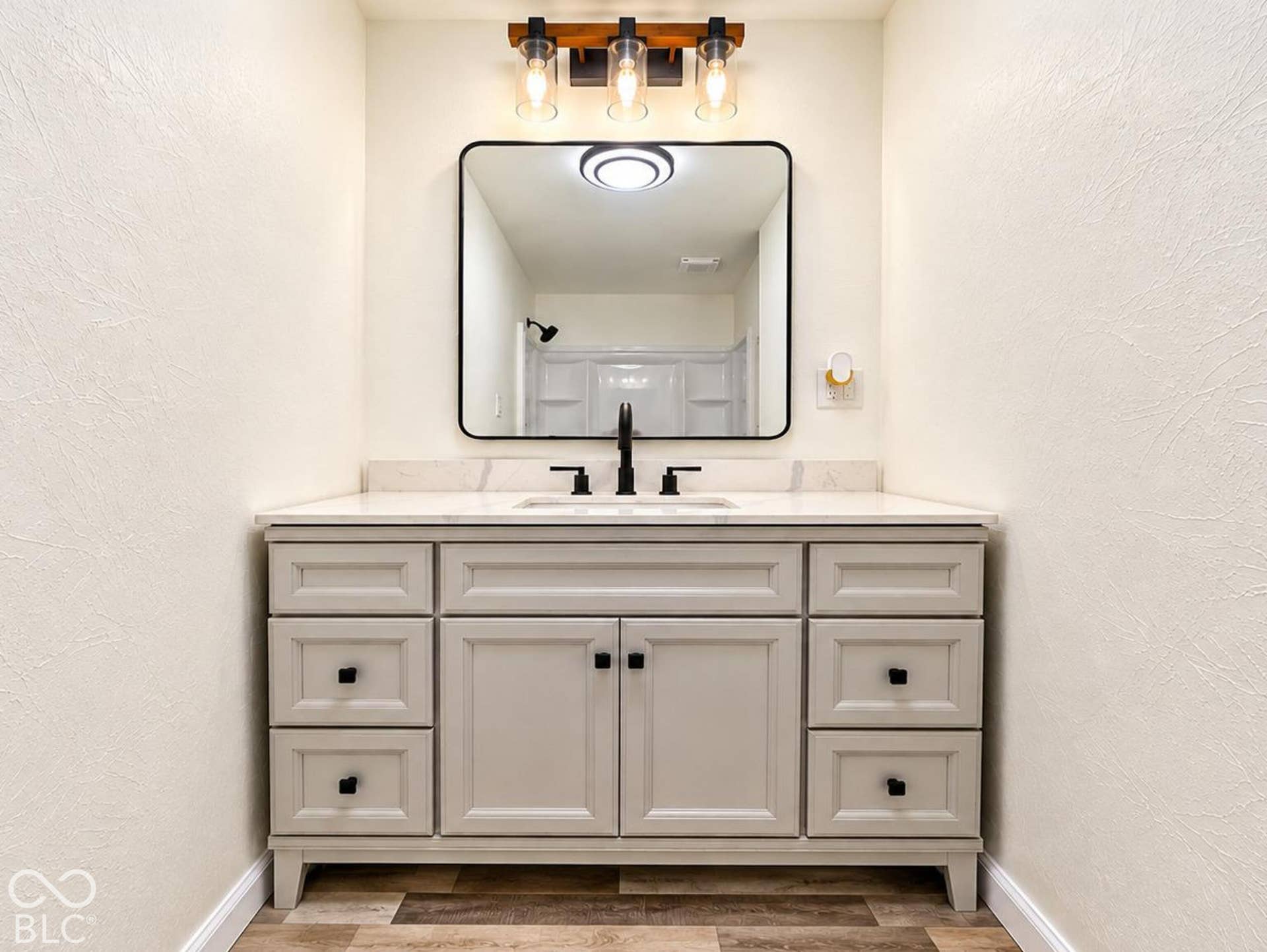 Vanity featuring a light gray finish, solid-surface countertop, matte black hardware, rectangular mirror with a black frame, and a three-light fixture with a wood-tone mount