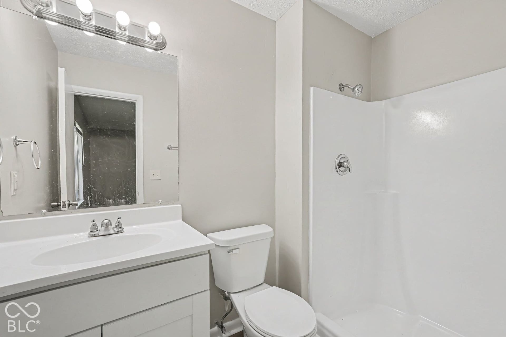 Bathroom featuring a white vanity with an integrated sink, a full-width vanity mirror, a four-bulb light fixture, and a white shower surround with chrome hardware