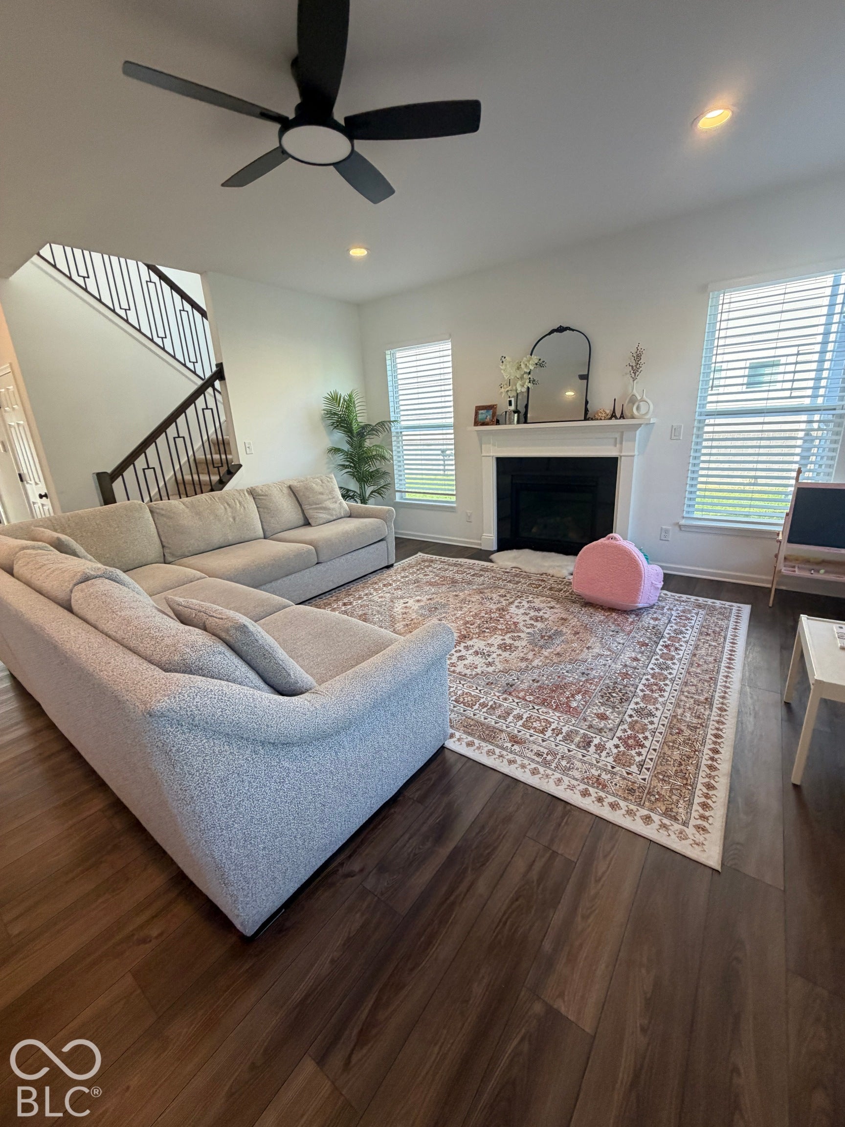 Living area featuring dark wood-finish flooring, a fireplace with a white mantel, recessed lighting, a modern ceiling fan, and a staircase with metal railings