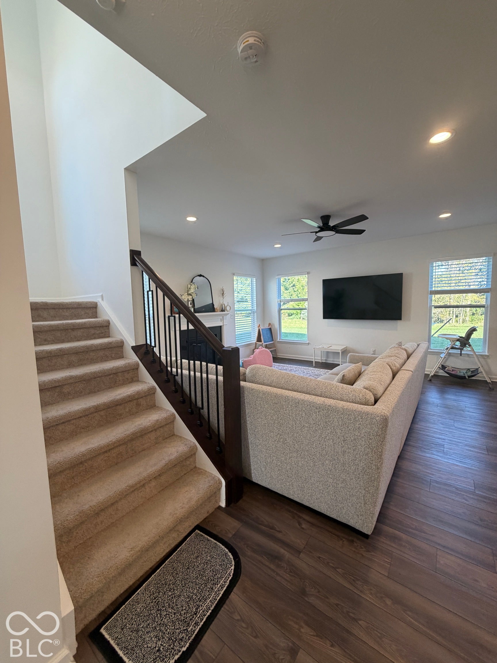 Carpeted staircase featuring a dark-finish wood handrail with vertical metal balusters, wood-finish flooring, recessed lighting, and a fireplace with a mantel