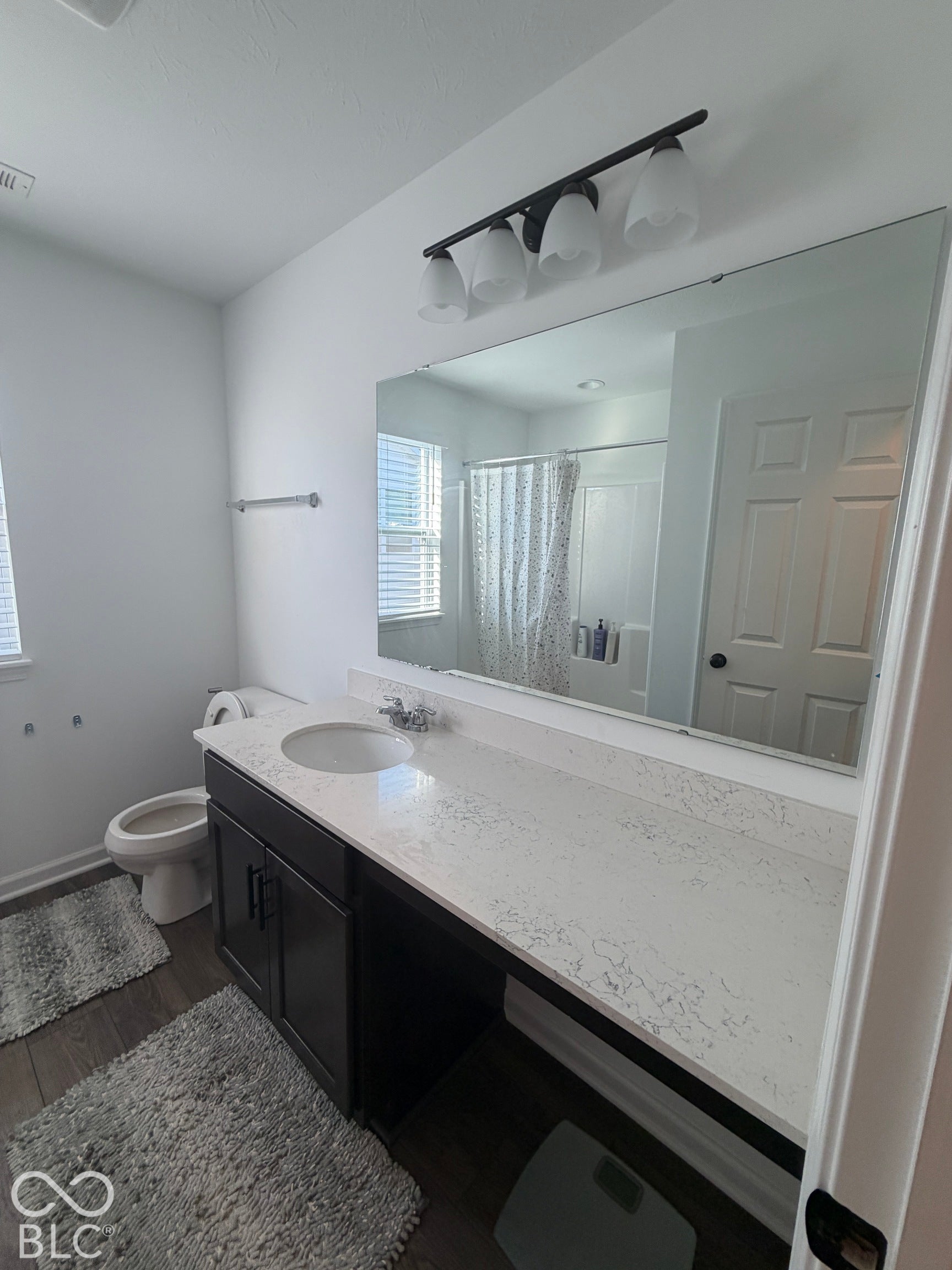 Bathroom featuring an extended vanity with a light-toned stone-finish countertop, dark-toned cabinetry, a full-width wall mirror, and a four-light fixture