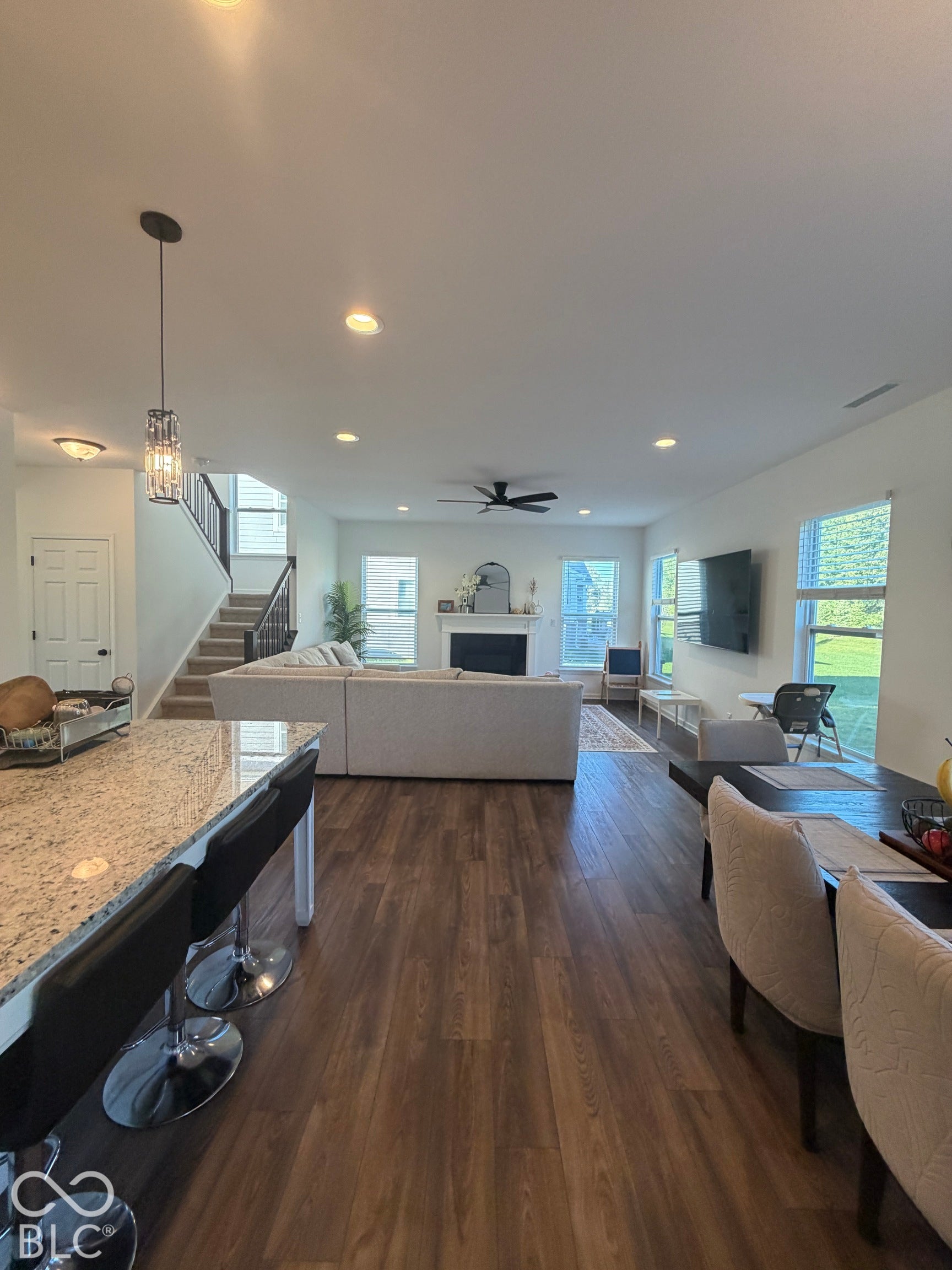 Open-concept interior featuring wood-finish flooring, stone-surfaced kitchen island, recessed lighting, fireplace with a mantle, and multiple oversized windows