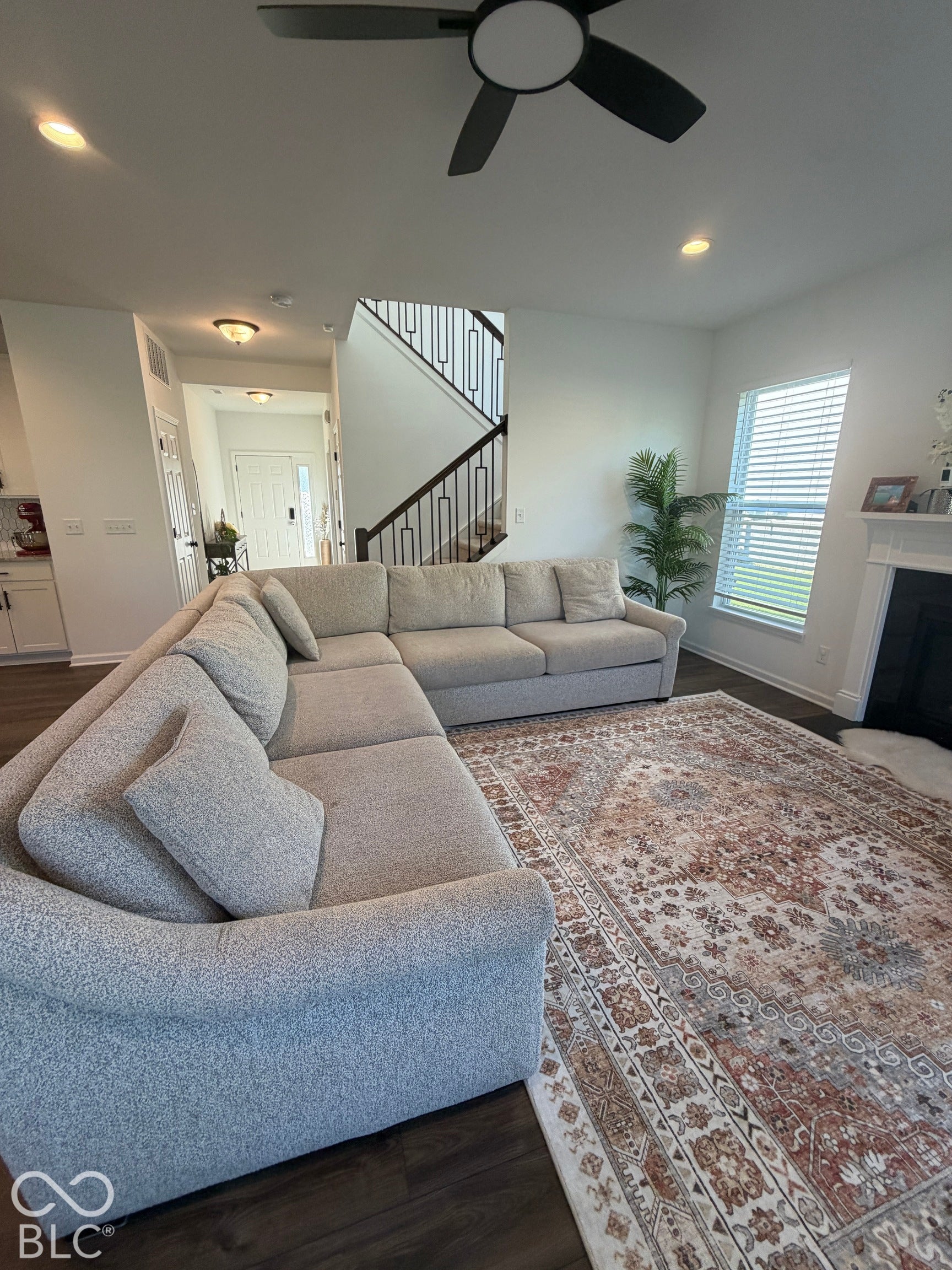 Open-concept living space featuring wood-finish flooring, recessed lighting, matte black ceiling fan with integrated lighting, staircase with iron railing, and fireplace with a decorative mantel