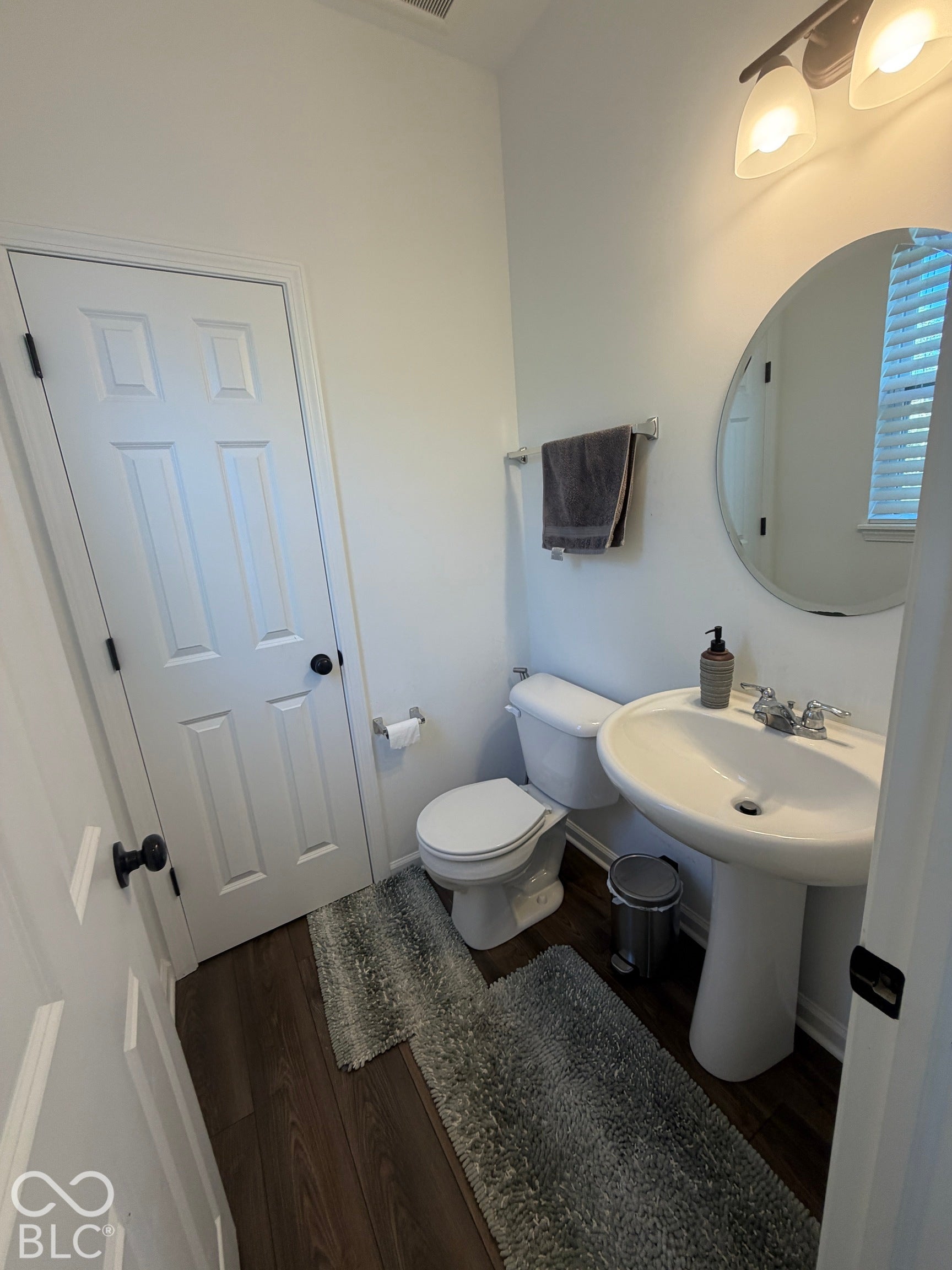 Powder room featuring wood-finish flooring, a white pedestal sink, chrome hardware, a circular wall-mounted mirror, and multi-light vanity fixture