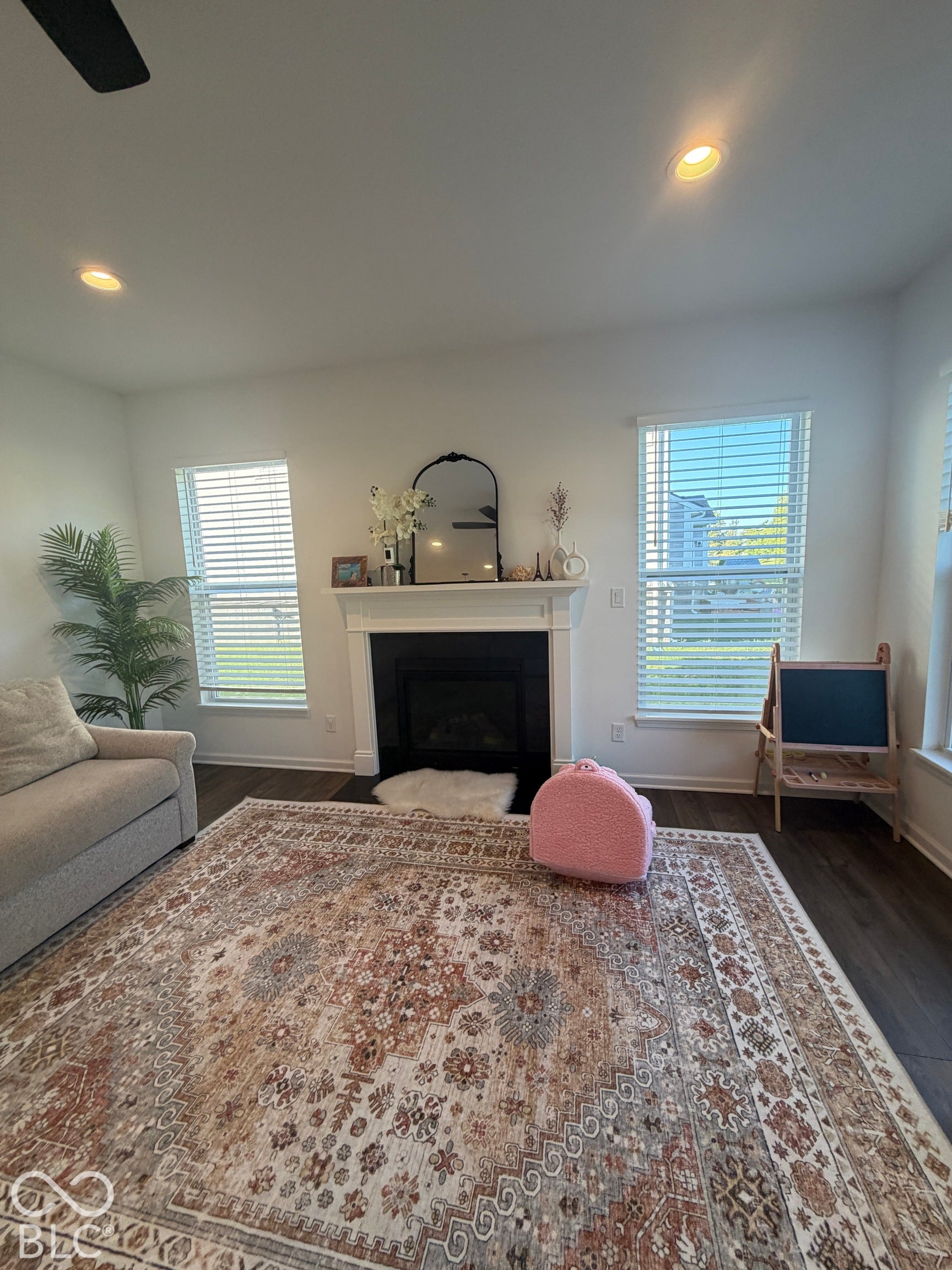 Fireplace with a classic white mantel and dark tile surround, flanked by windows with horizontal blinds, complemented by recessed lighting and wood-finish flooring