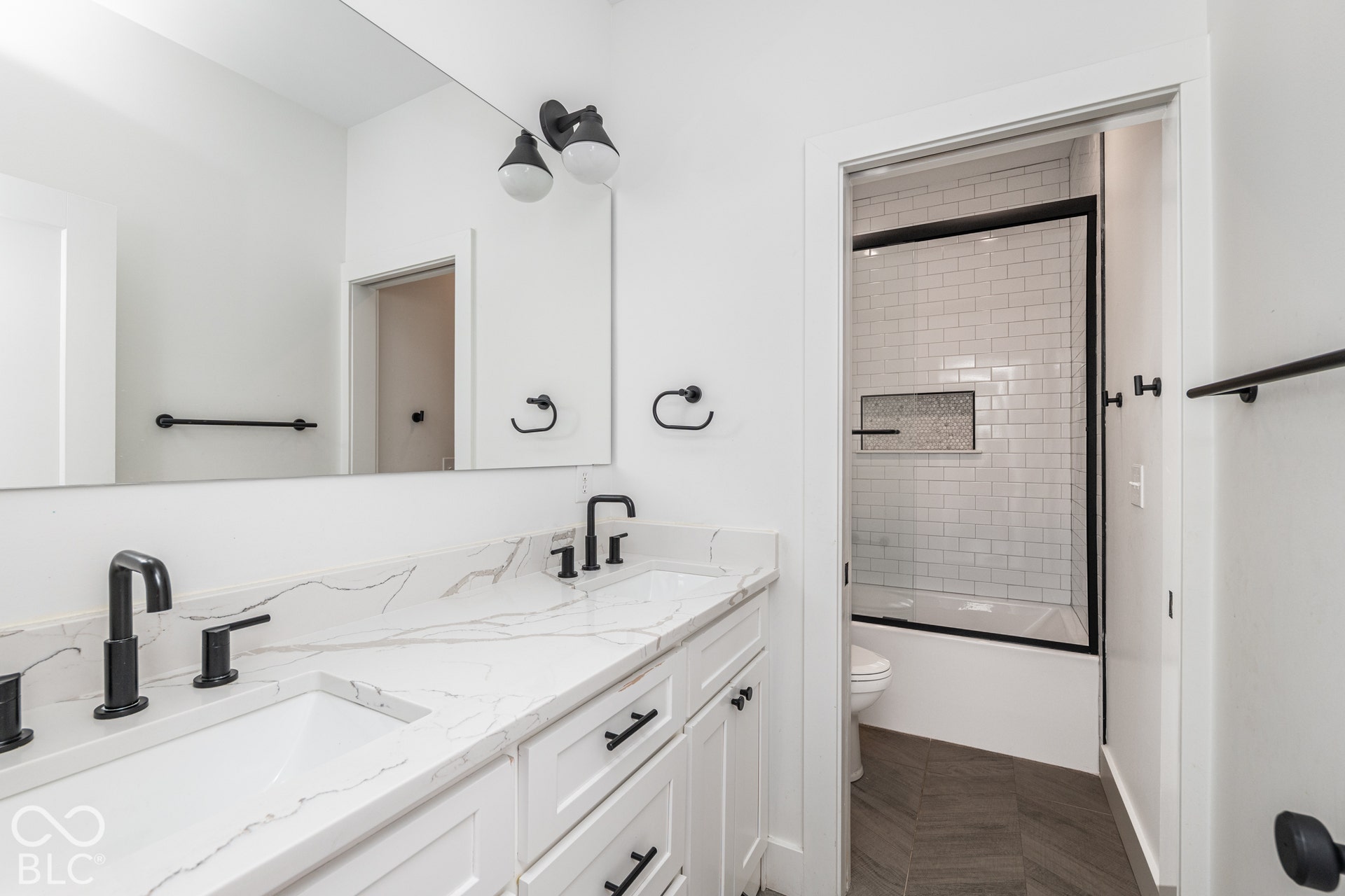 1401 Olive Street, Indianapolis, IN 46203 Double vanity featuring stone-finish countertops, matte black plumbing fixtures, white shaker-style cabinetry, a full-width mirror, and a tiled shower surround with a built-in niche