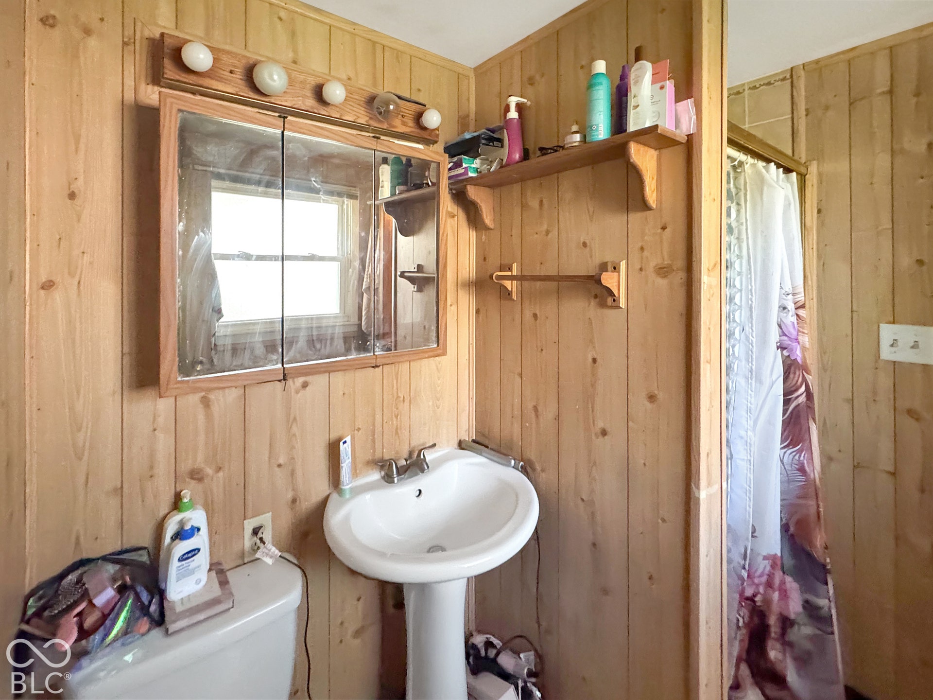 Wood-paneled bathroom featuring a pedestal sink, triple-panel medicine cabinet, wall-mounted light fixture, and wooden wall shelving