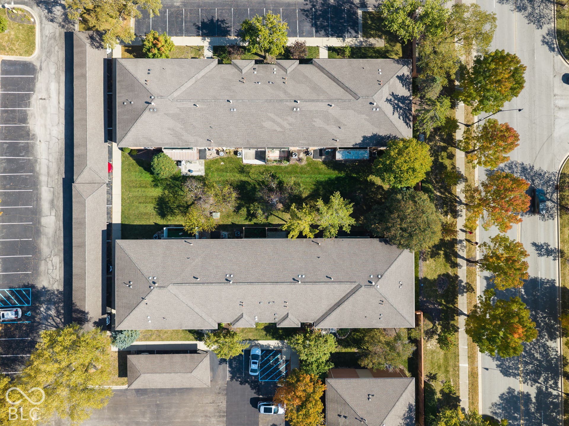 Aerial perspective of a multi-building residential property featuring shingled roofs, dedicated asphalt parking lots, mature trees, and landscaped green space