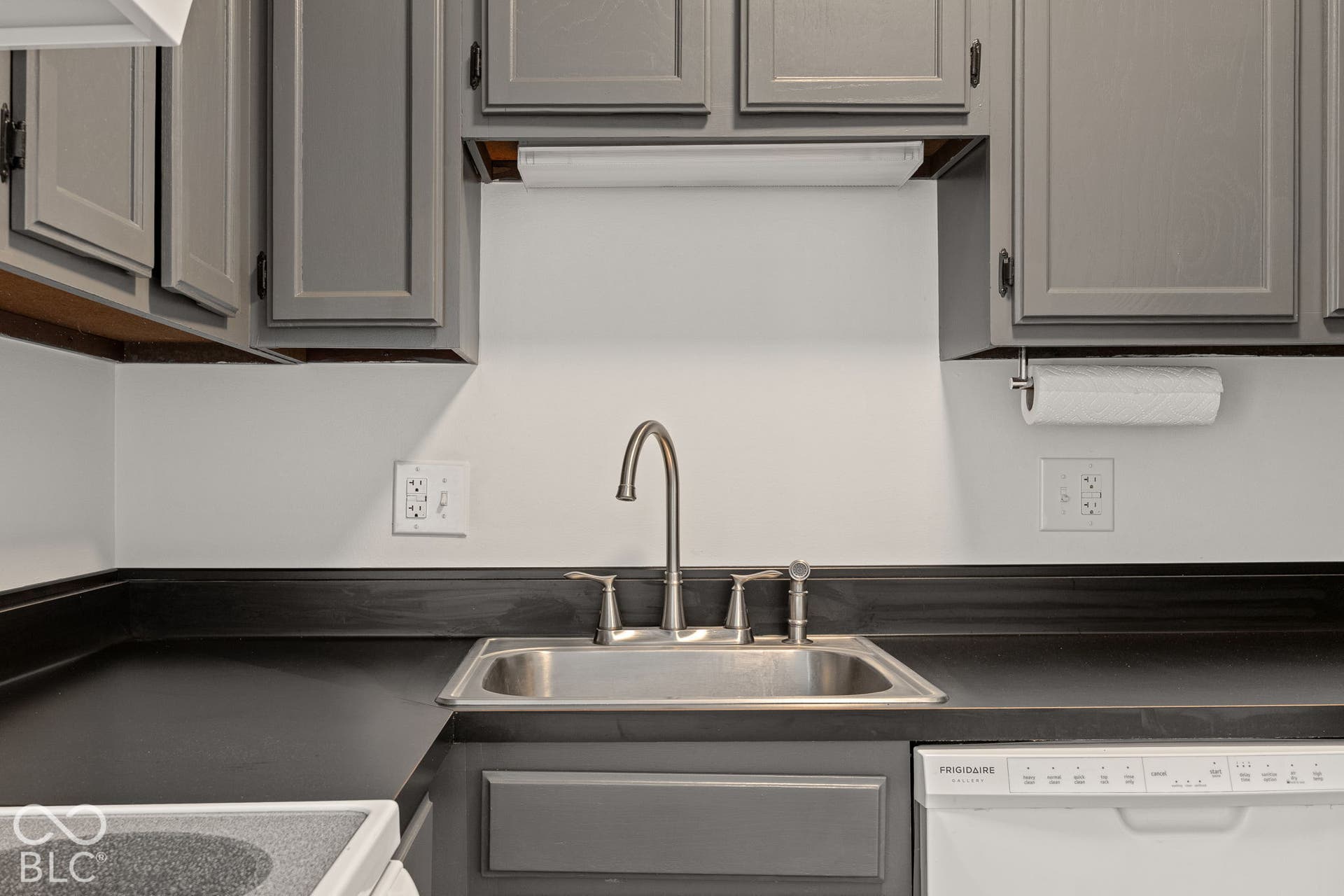 Kitchen workspace featuring grey shaker-style cabinetry, matte black countertops with integrated backsplash, a stainless steel undermount sink with high-arc gooseneck faucet, and a built-in dishwasher