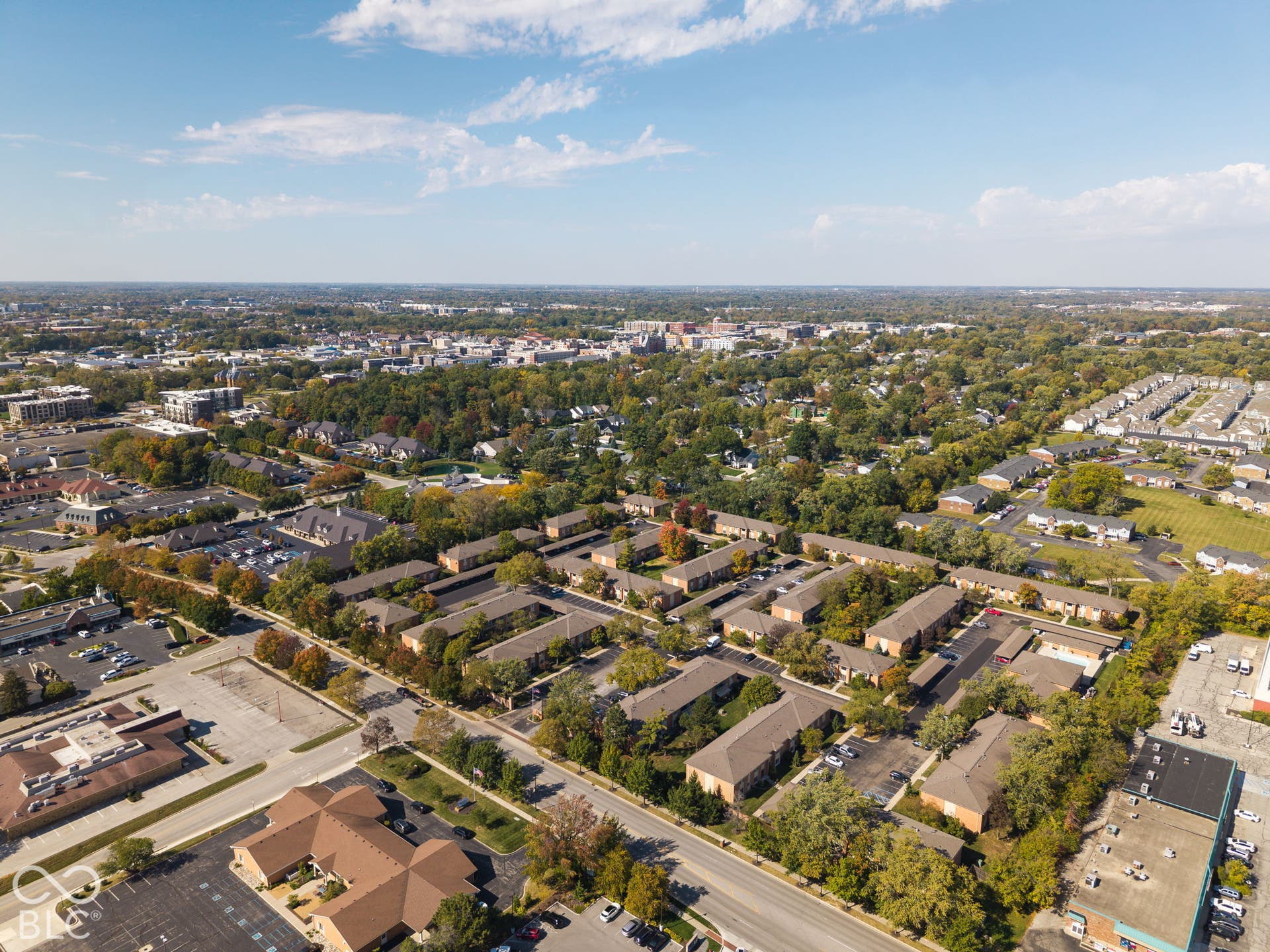 Aerial perspective featuring a multi-building property complex with asphalt paving, concrete walkways, mature trees, and shingled rooflines