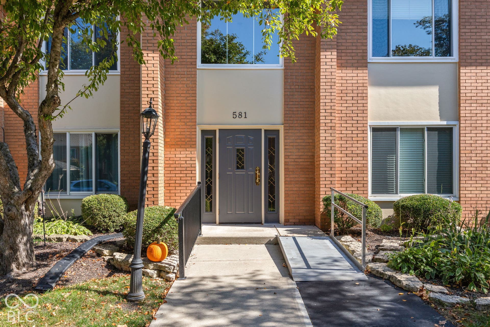 Multi-story exterior featuring tan brick masonry, a central entryway with a paneled door, decorative sidelights, a metal accessibility ramp, and stone-bordered landscaping