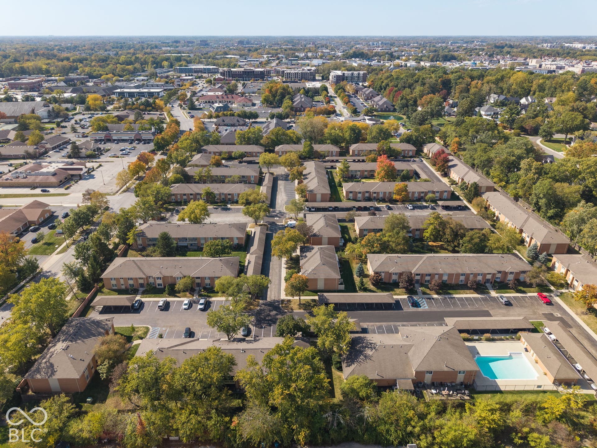 Aerial perspective of multi-building property featuring brick-exterior structures, asphalt-paved parking areas, covered carport stations, and a fenced outdoor swimming pool