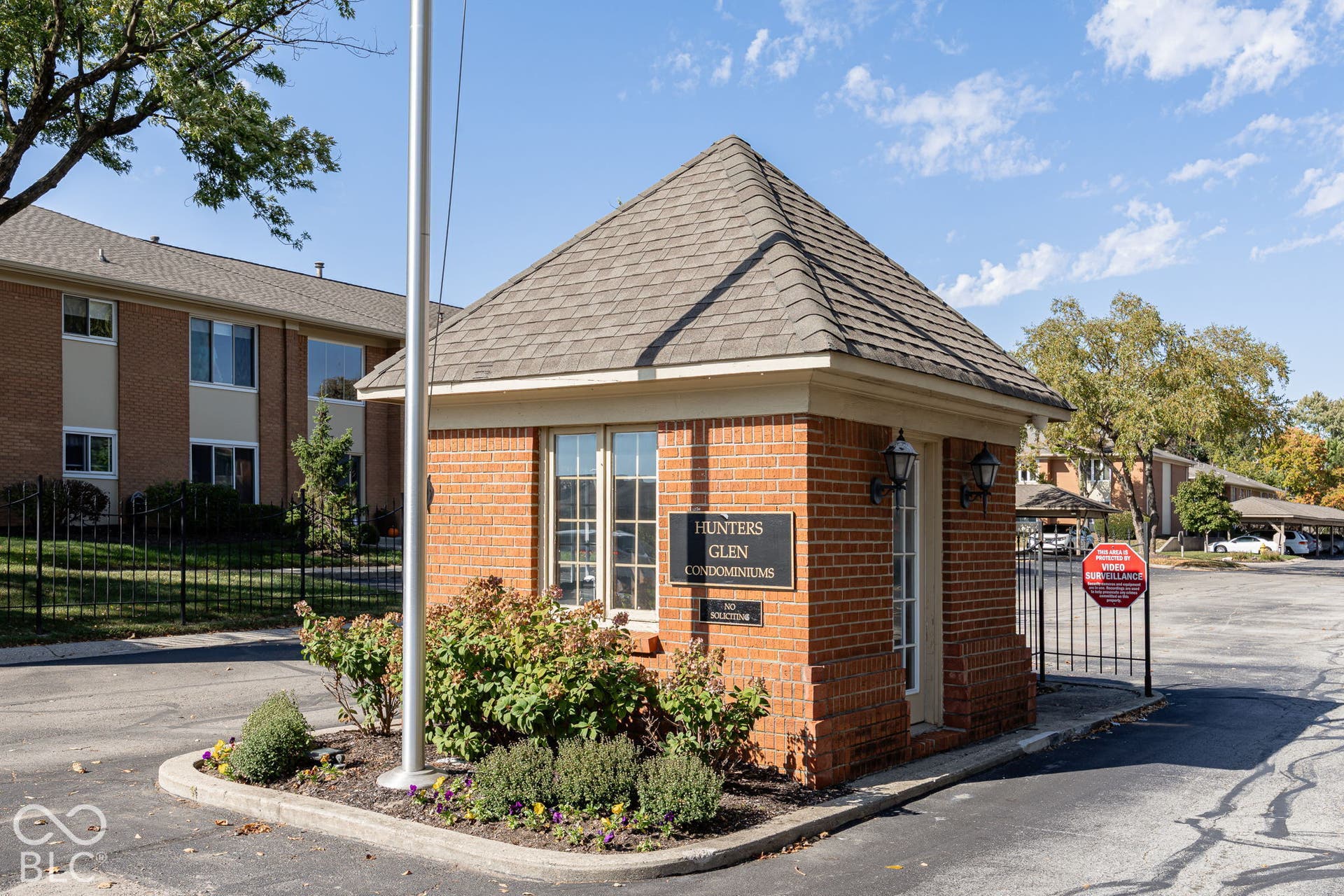 Brick gatehouse entrance featuring a hipped shingle roof, iron perimeter fencing, and exterior wall-mounted lanterns