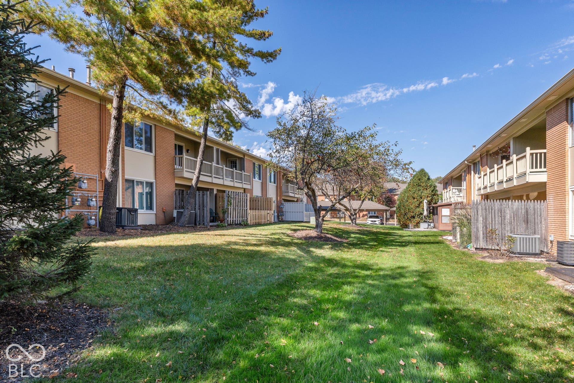 Brick exterior multi-unit building featuring upper-level balconies, ground-level privacy partitions, mature trees, and expansive lawn space