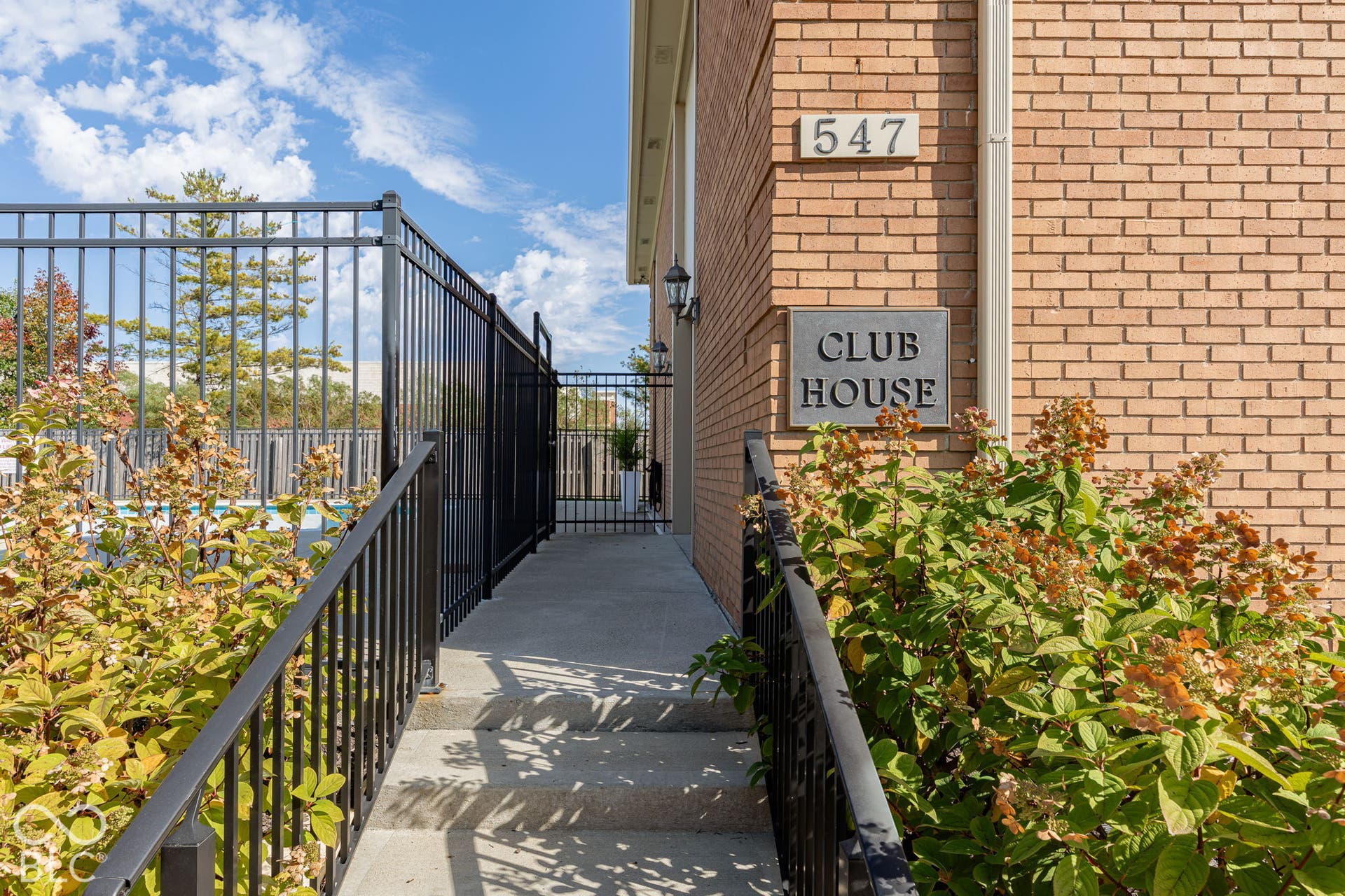 Exterior brick clubhouse building featuring a concrete walkway, black metal fencing, and decorative exterior wall-mounted lighting