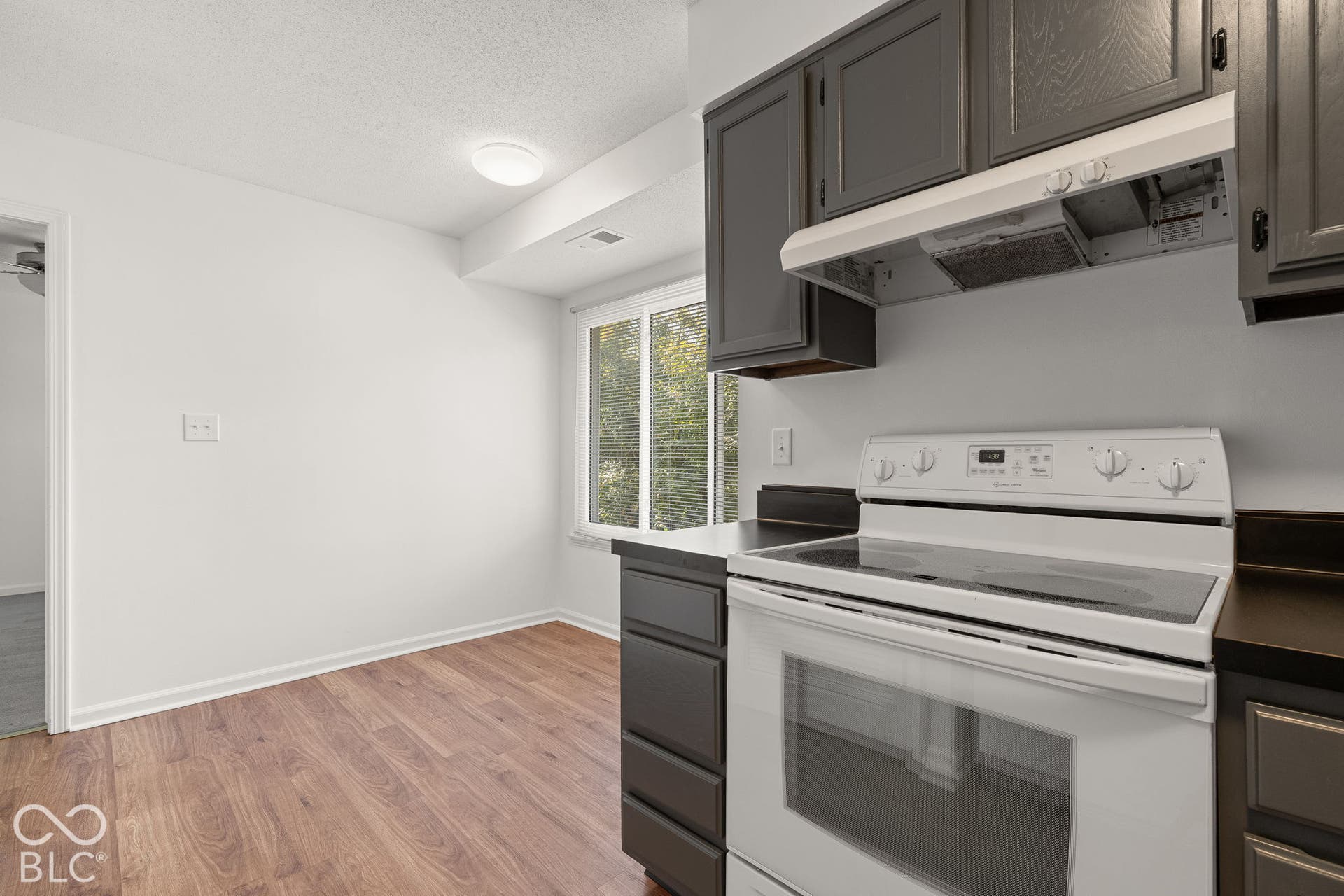 Kitchen area featuring dark-toned cabinetry, a white electric range, an integrated range hood, a sliding window, and wood-finish flooring