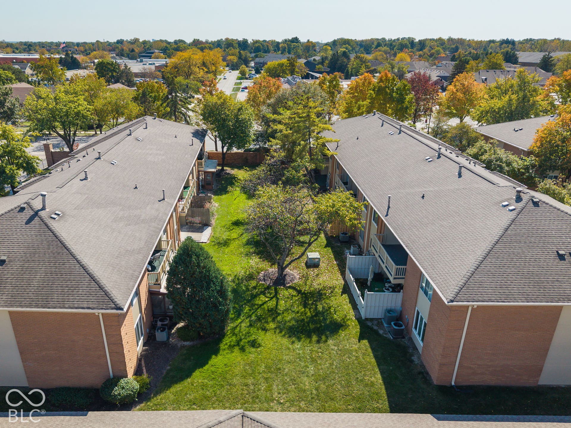 Aerial perspective of two multi-family residential structures featuring brick veneer facades, shingled roofing, private balconies, and an expansive common green space with mature trees