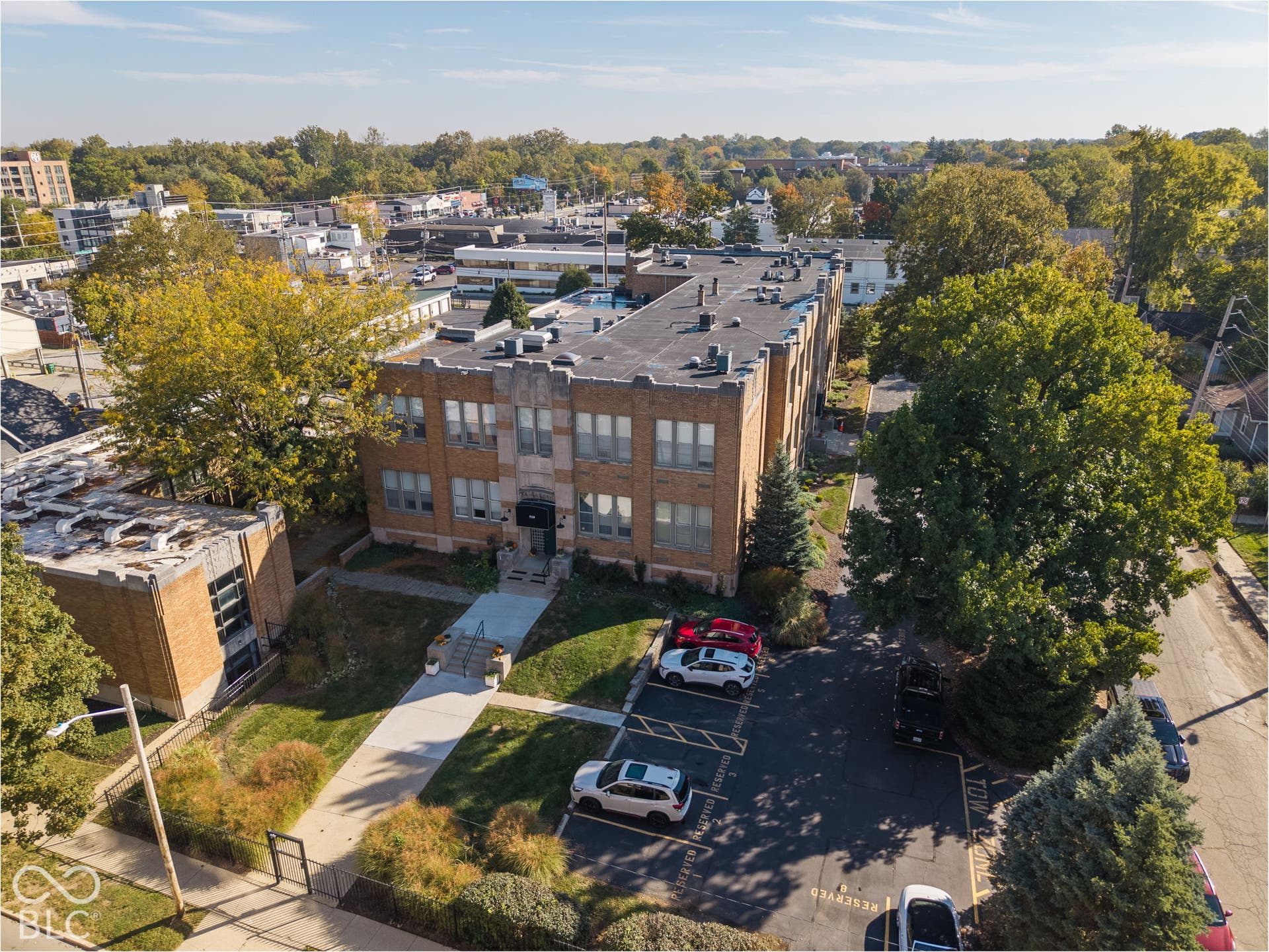 Multi-story brick building exterior featuring a flat roofline, paved parking area, and landscaped grounds with mature trees