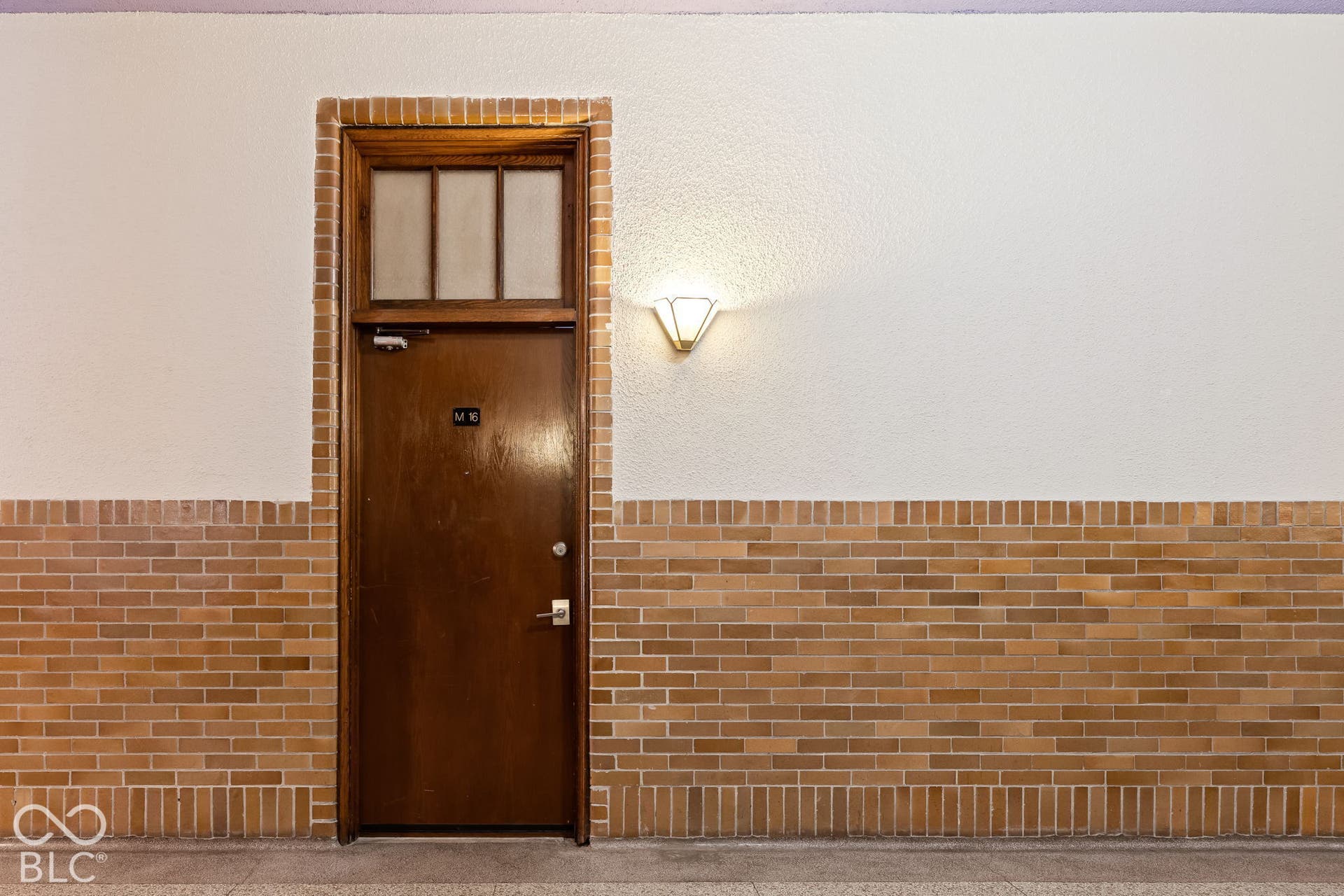 Solid wood door with a glass transom, framed by exposed brick masonry and featuring a classic wall-mounted sconce