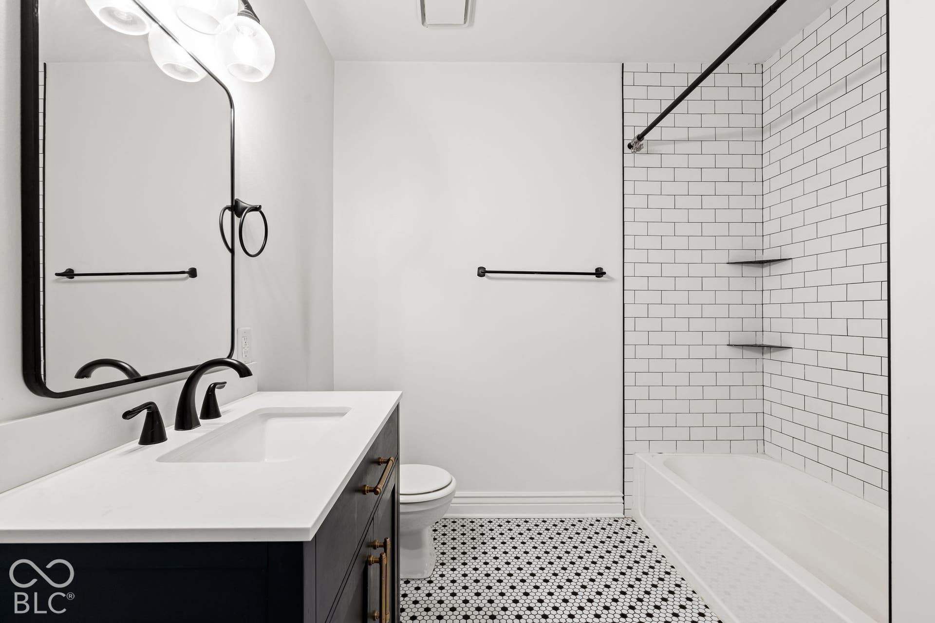 Dark-finish vanity featuring a white surface, undermount sink, and matte black hardware, paired with a subway-tiled tub surround, integrated shelving, and geometric-patterned flooring