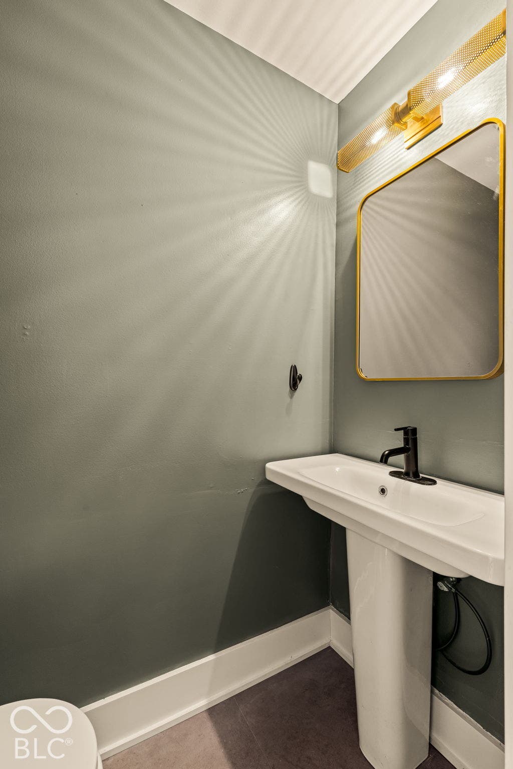 Pedestal sink with a matte black single-handle faucet, rectangular mirror with gold-tone frame, and wall-mounted gold-tone vanity light fixture