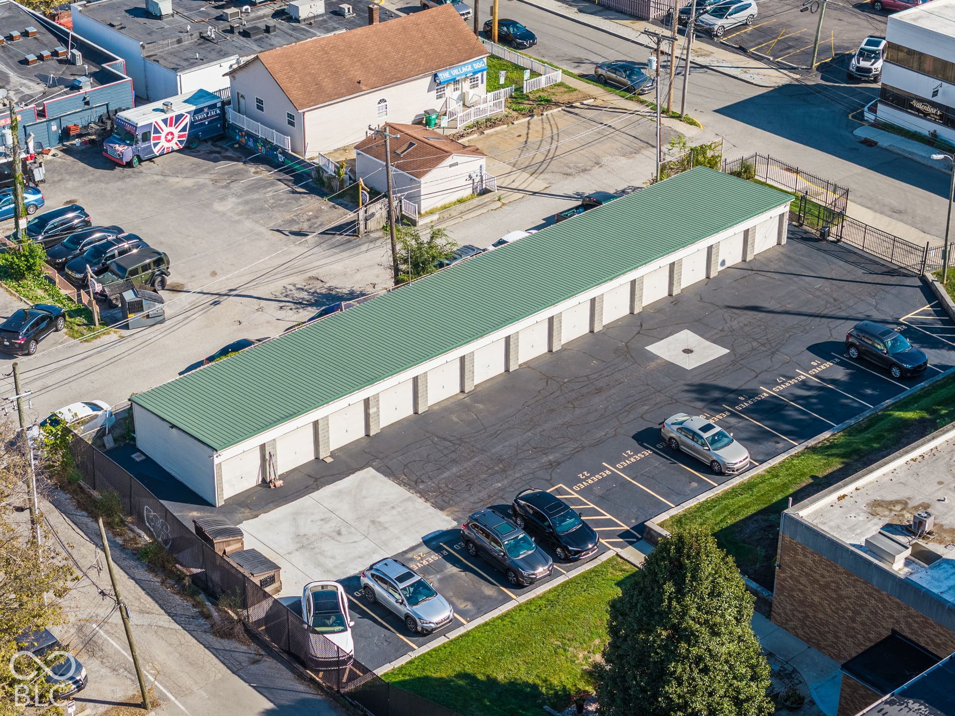 Aerial perspective of a multi-unit storage facility featuring a green metal roof, individual roll-up doors, and an asphalt parking area with demarcated parking stalls